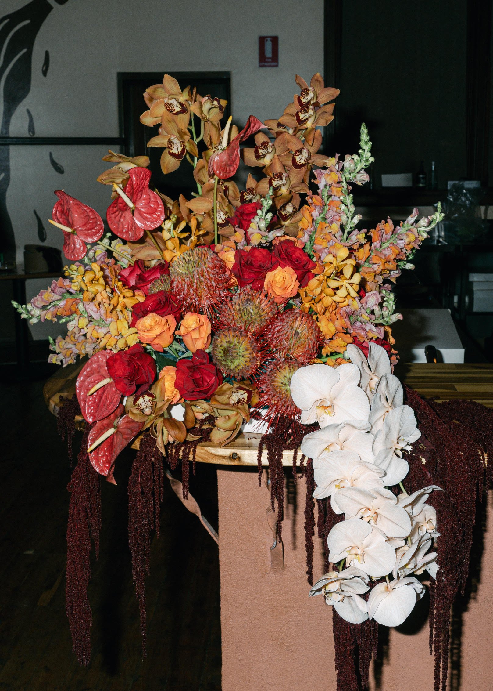 A large floral arrangement on a wooden pedestal, featuring orchids, roses, and other tropical flowers in shades of red, orange, pink, and white, with dark hanging beads.
