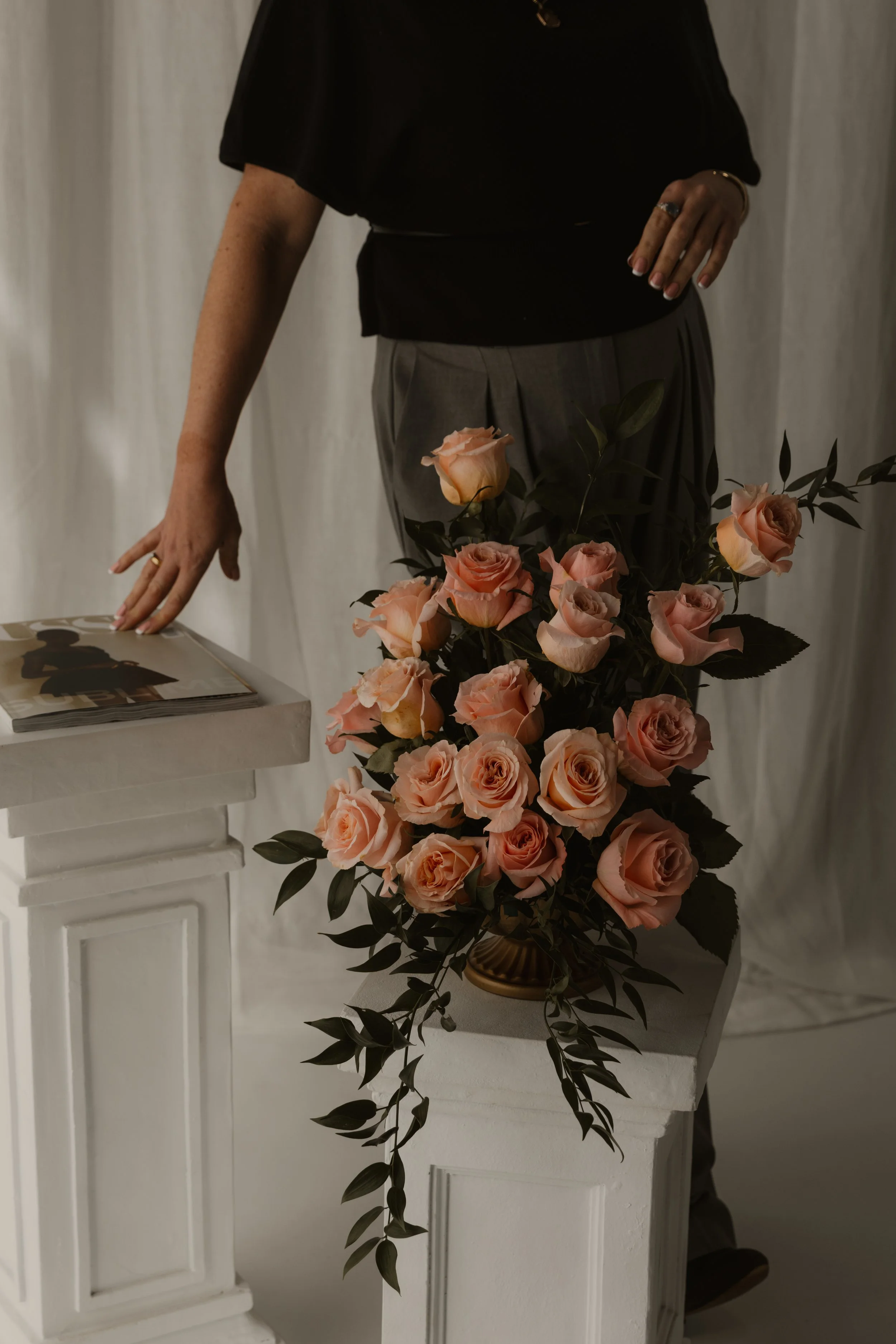 A person dressed in a black top and gray pants, standing next to a white table with a glossy magazine, with a large bouquet of pink roses and green foliage in a gold vase in front of them. The person is wearing rings and a bracelet.