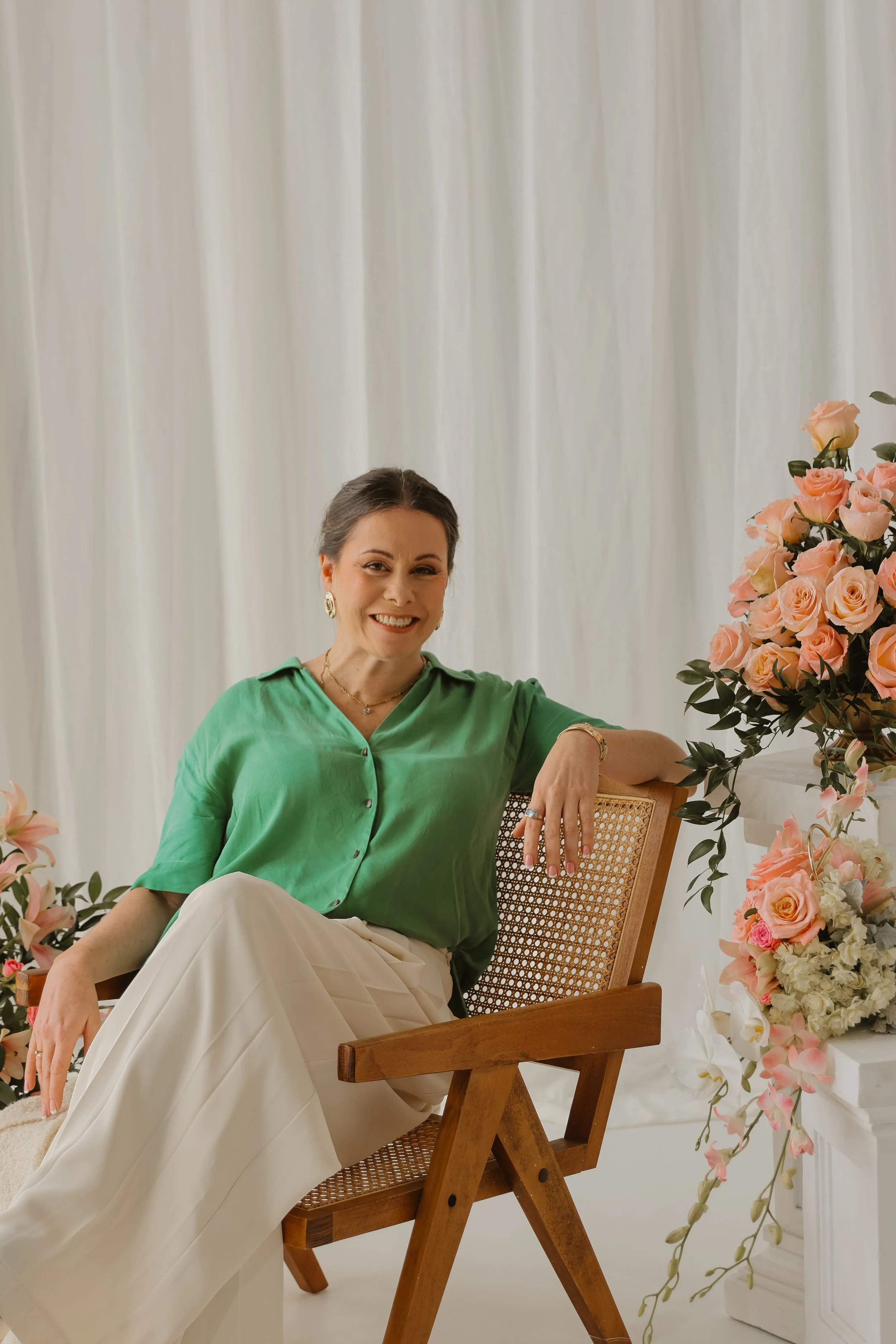 A woman with dark hair and earrings sitting in a wooden chair, smiling, surrounded by pink and white flowers, wearing a green shirt and cream-colored pants, in a bright room with white curtains.