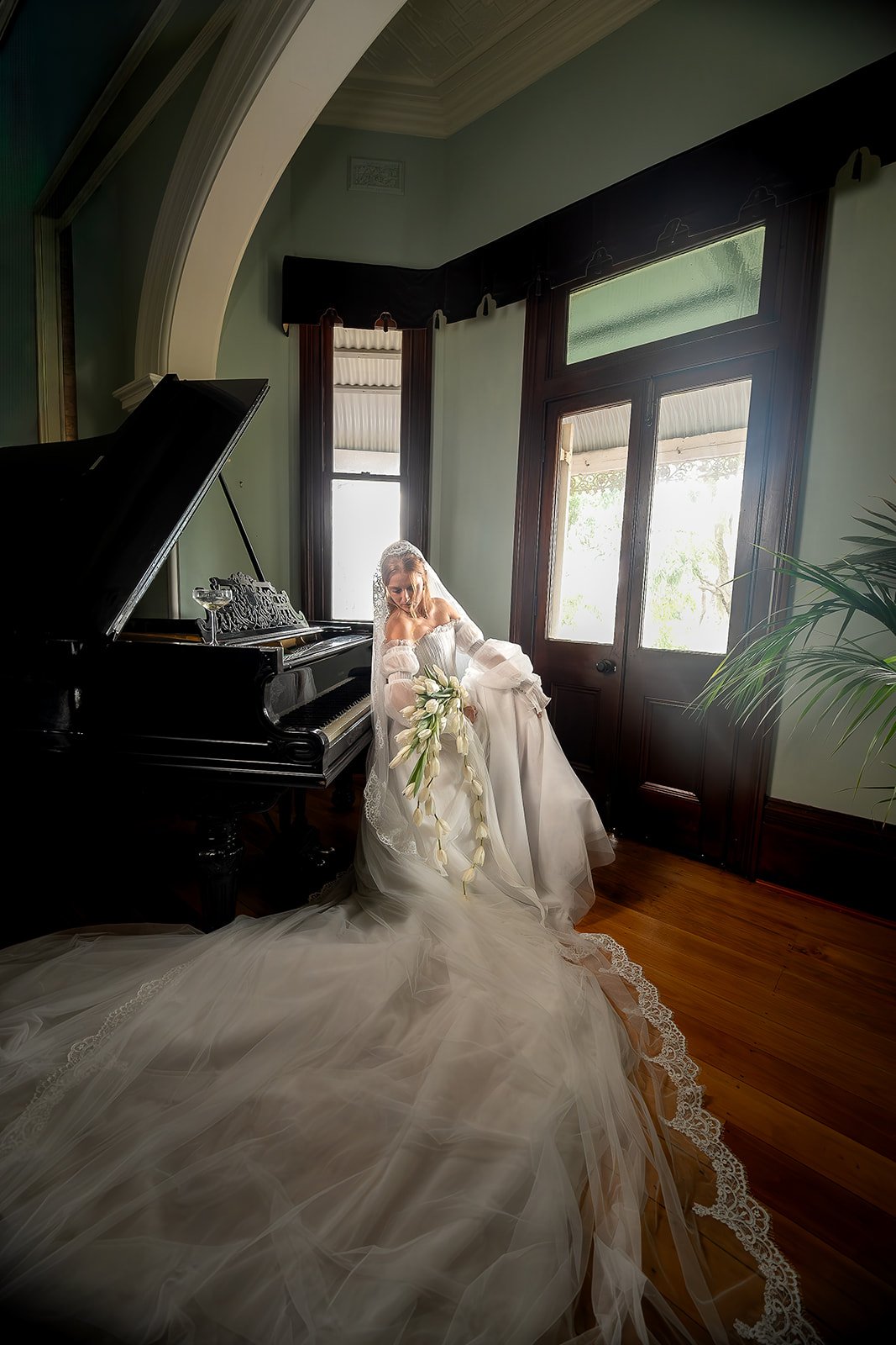 A bride in a white wedding gown is standing next to a black grand piano in a room with large windows and dark wood trim. She is holding a bouquet of white flowers and looking down, with natural light coming through the windows.