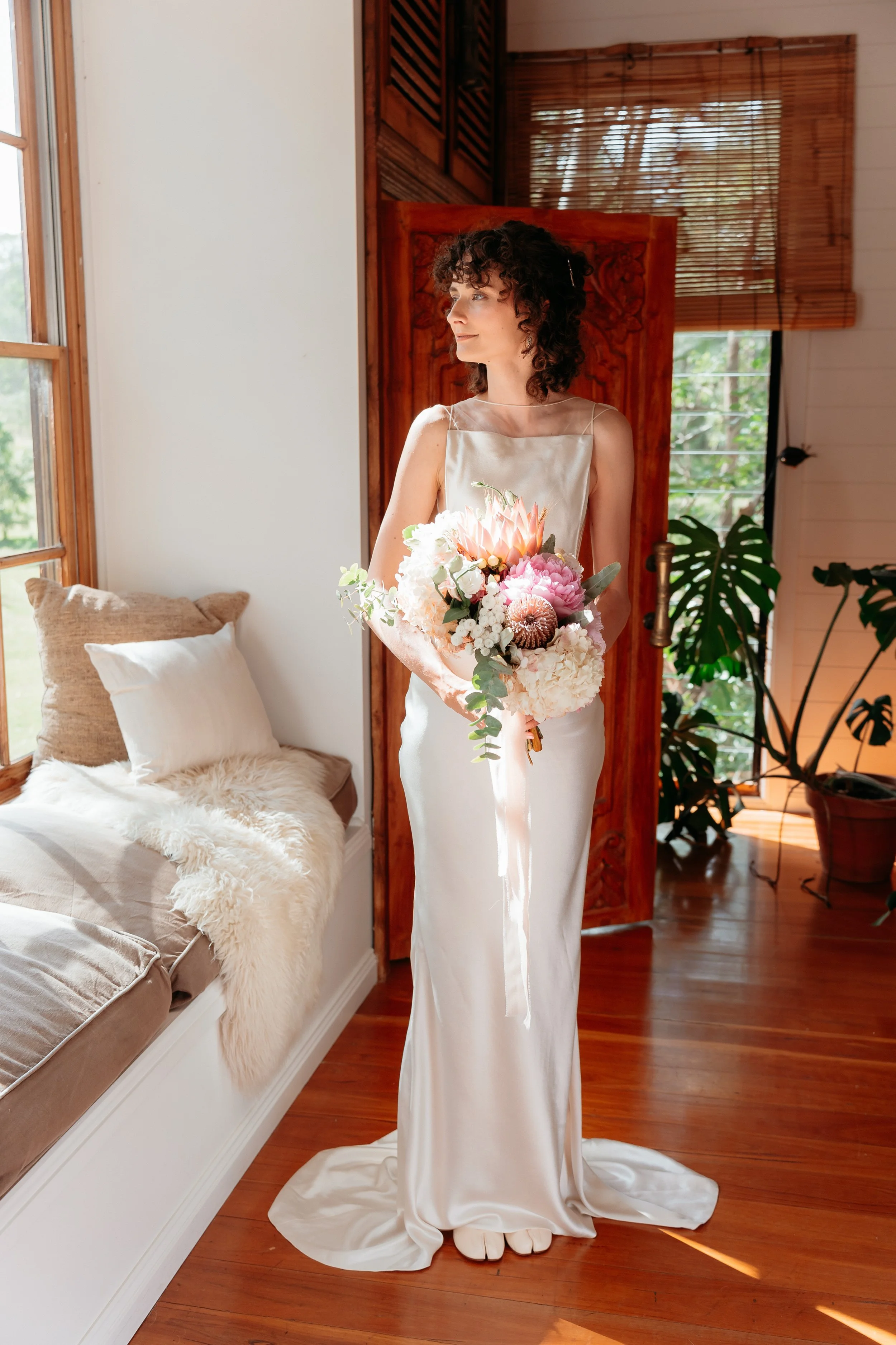 A woman in a white dress holding a bouquet of flowers standing inside a room near a window with bamboo blinds. The room has wooden flooring, a bed with pillows and a throw blanket, and plants near the window.