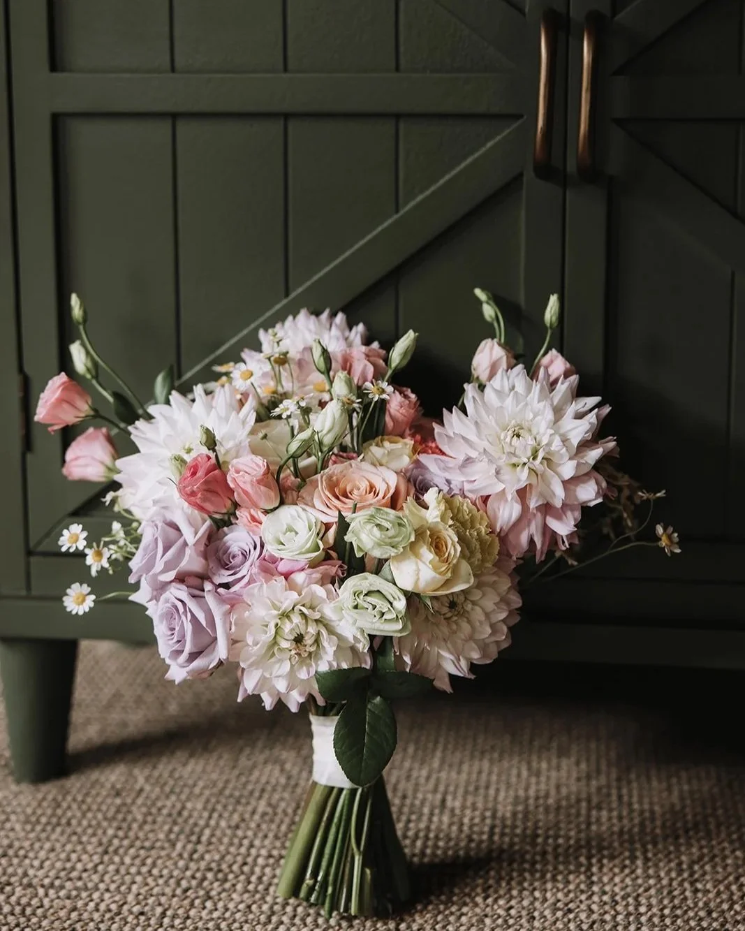 Bouquet of pink and white flowers, including roses, dahlias, and daisies, placed on a textured carpet in front of a dark green cabinet.