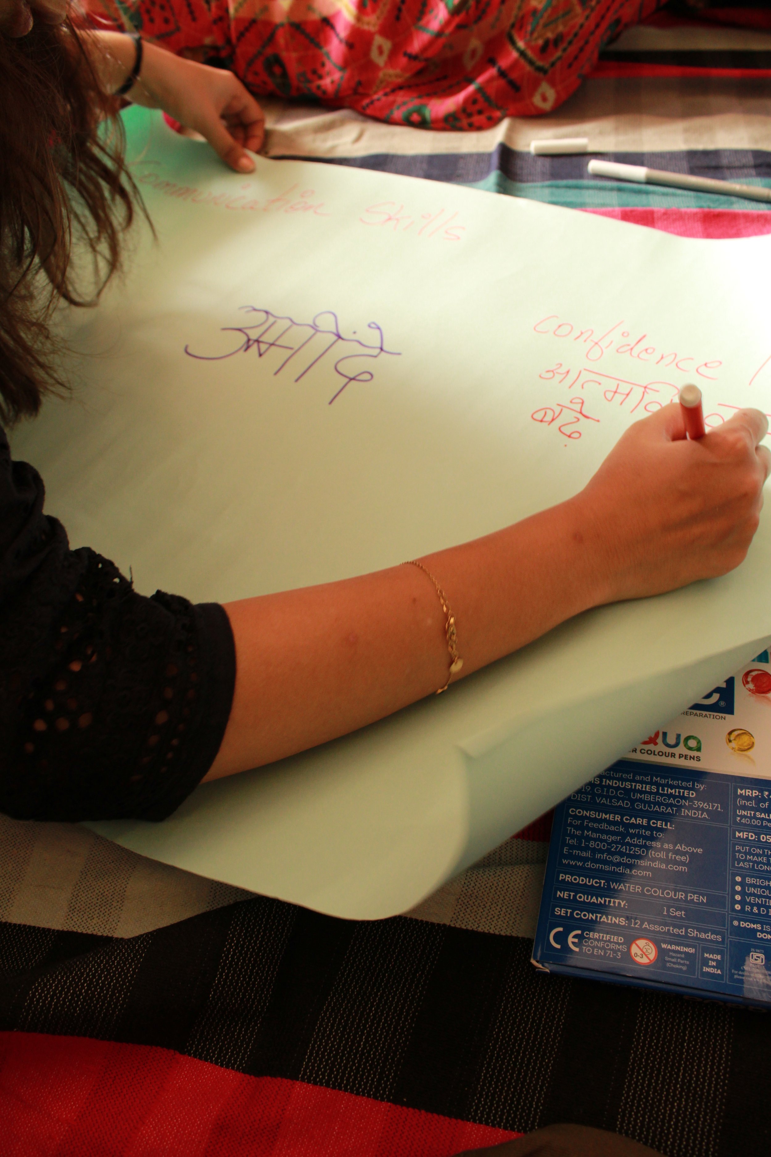 A person with long, curly hair is writing on a large sheet of white paper with a red marker. The paper is on a table with a multicolored cloth. There are two white markers nearby and a package of water color pens on the side.