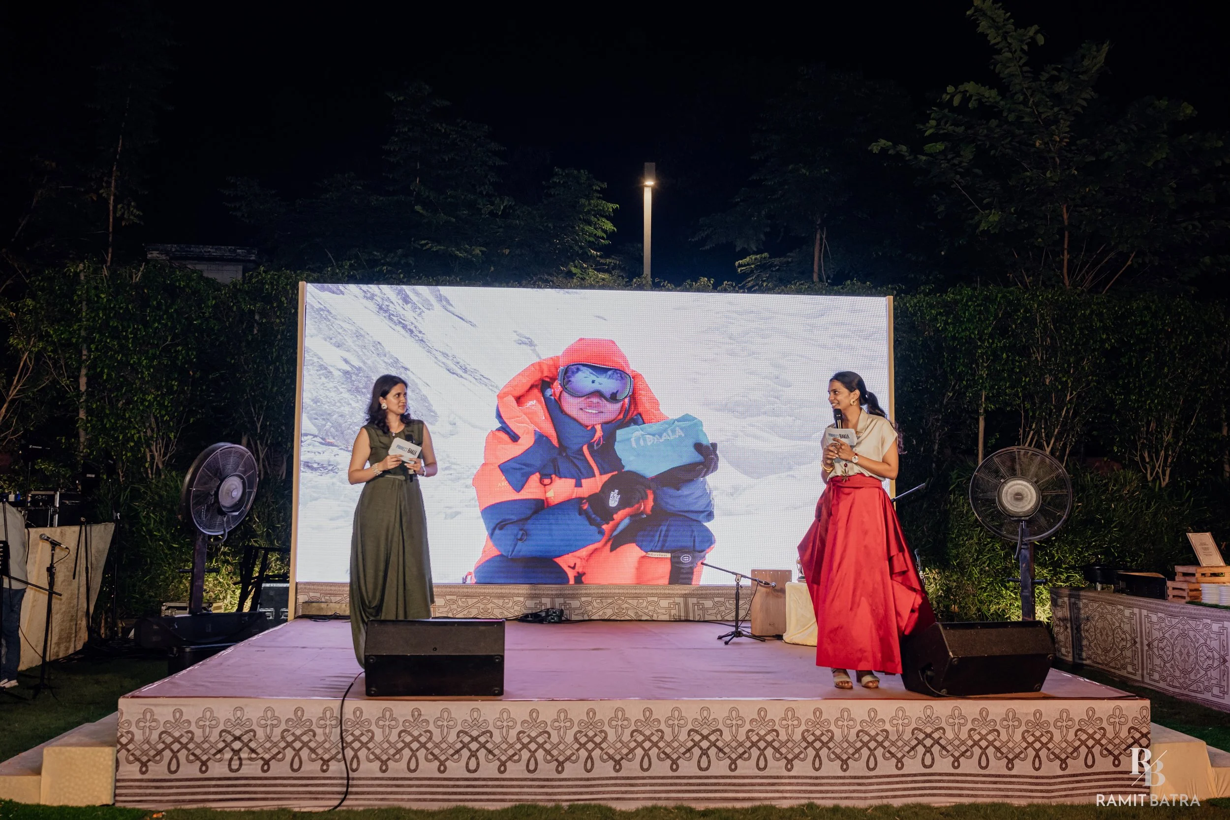 Two women are standing on a stage outdoors, having a conversation. Behind them is a large LED screen displaying an image of a child in winter gear. The setting is nighttime, with surrounding trees and outdoor fans visible.