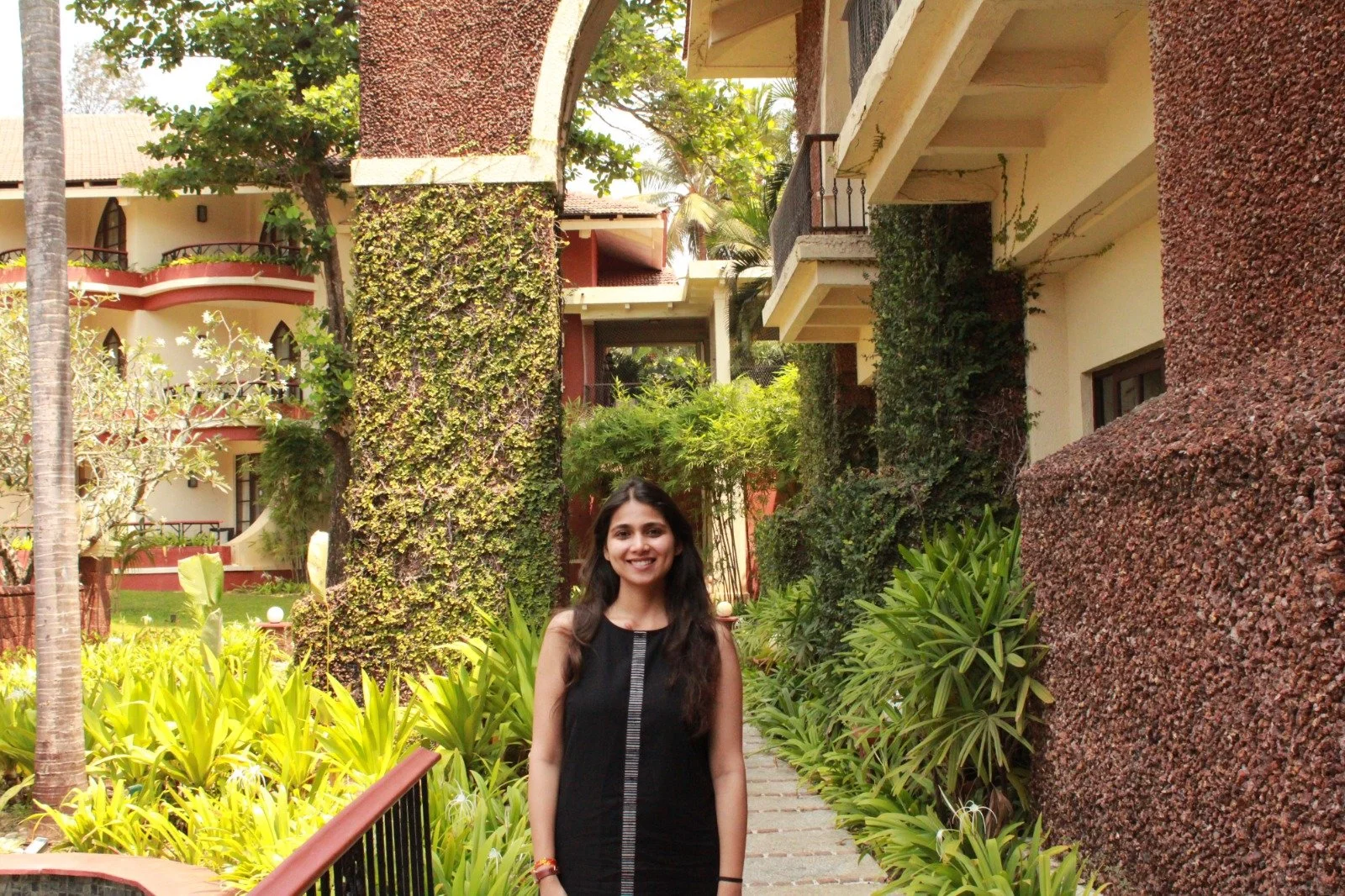 A smiling woman in a black dress standing on a garden pathway surrounded by lush green plants and tall decorative walls within a courtyard of a building complex.