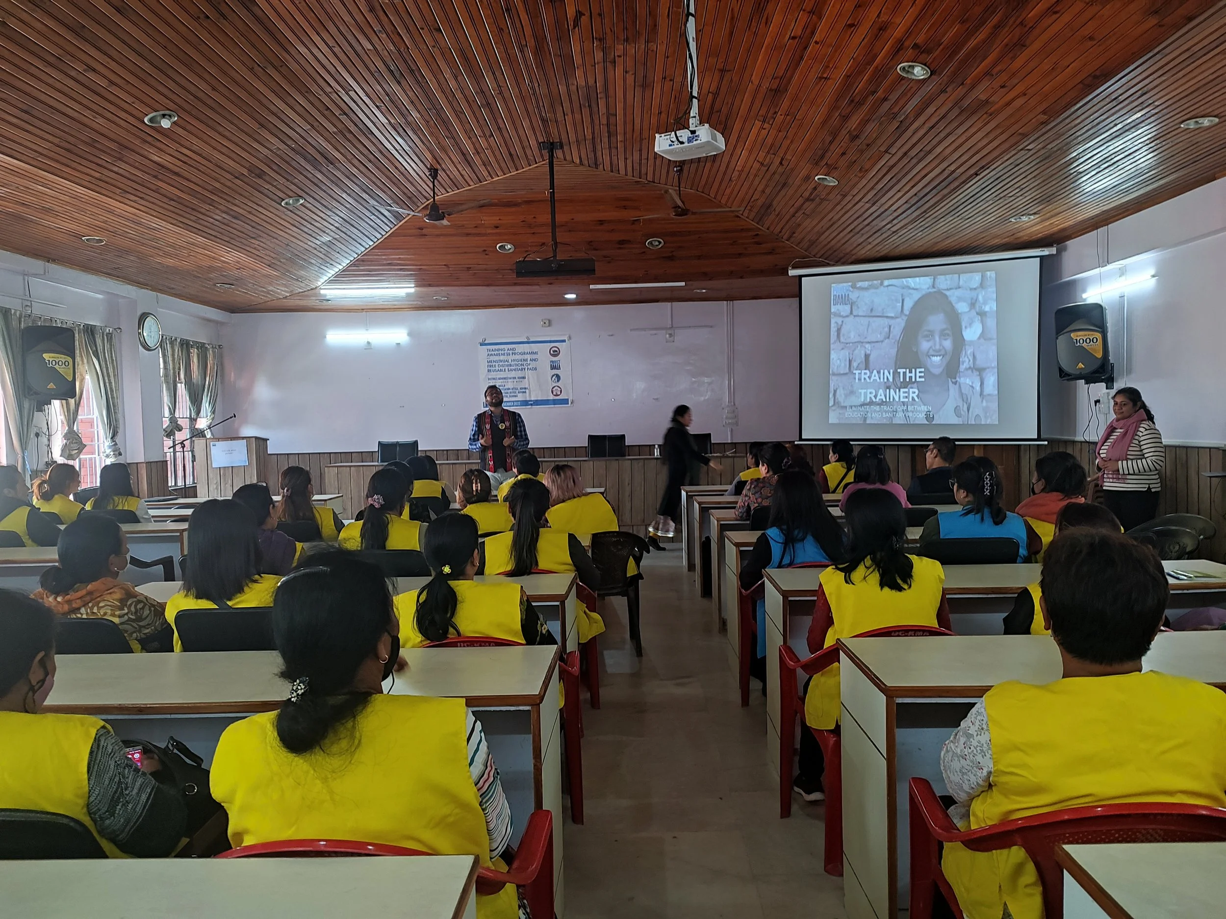A classroom with many students wearing yellow vests, seated and facing a speaker and a large screen displaying a person smiling, with the words 'Train the Trainer'.