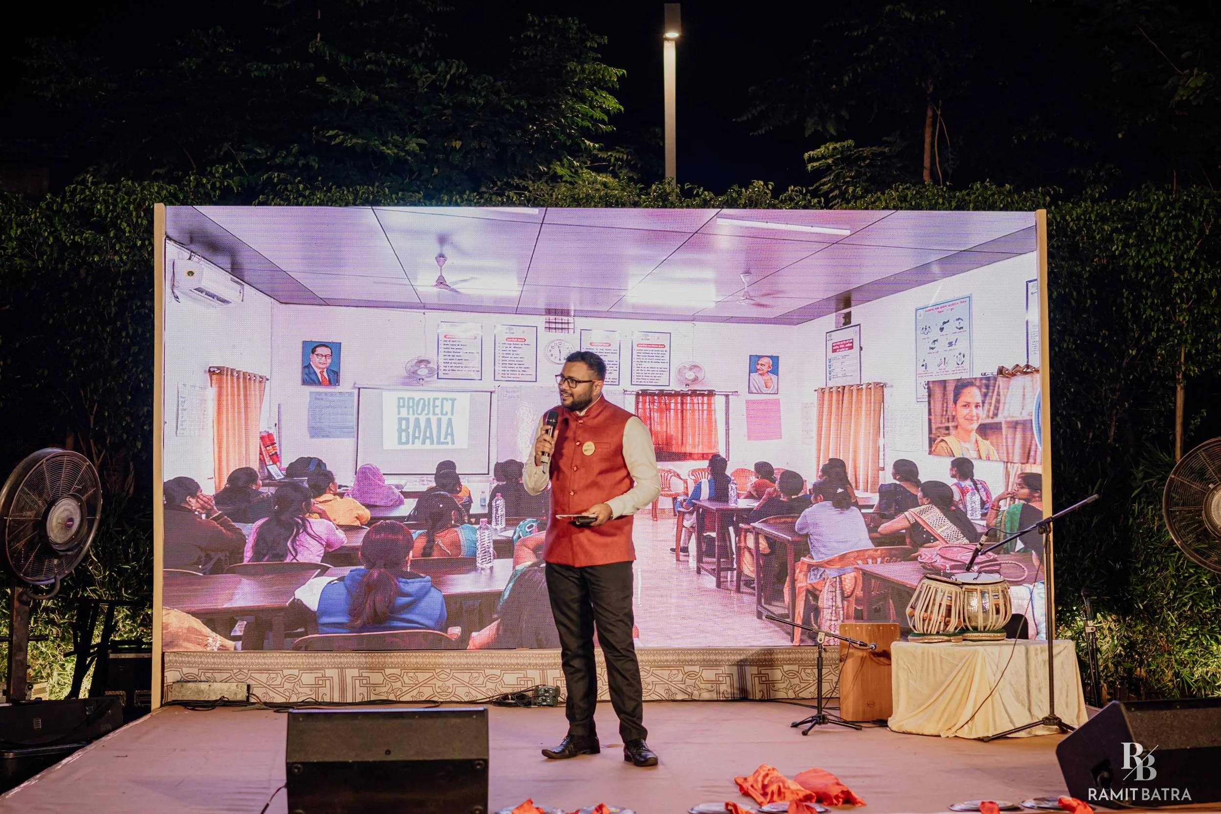 A man in a maroon vest and glasses stands on a stage holding a microphone and a tablet. Behind him is a large screen showing an indoor scene with people sitting at tables, a bulletin board, and portraits of historical figures. The stage is outdoors at night, surrounded by greenery and illuminated by a streetlamp.
