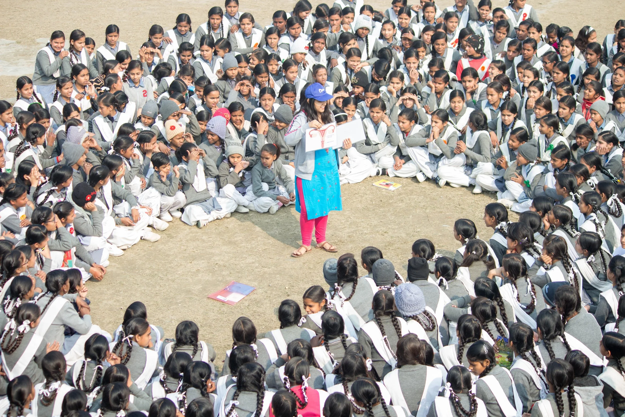 A large group of schoolgirls in gray uniforms sitting outdoors on the ground, attending a health education session where a woman is holding a diagram of the female reproductive system.