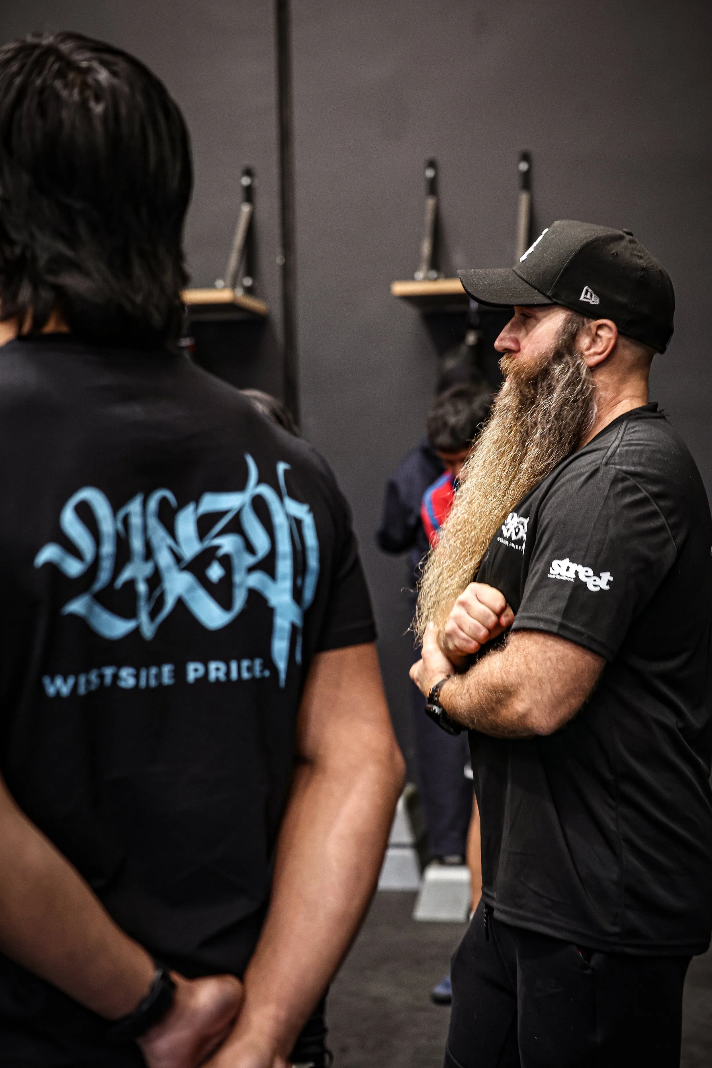 Two men are engaged in conversation, one with long black hair and wearing a black shirt with blue graffiti-style text, the other with a long, thick beard, wearing a black cap and black shirt with logos. The setting appears to be indoors with a dark background and shelves with tools or equipment.