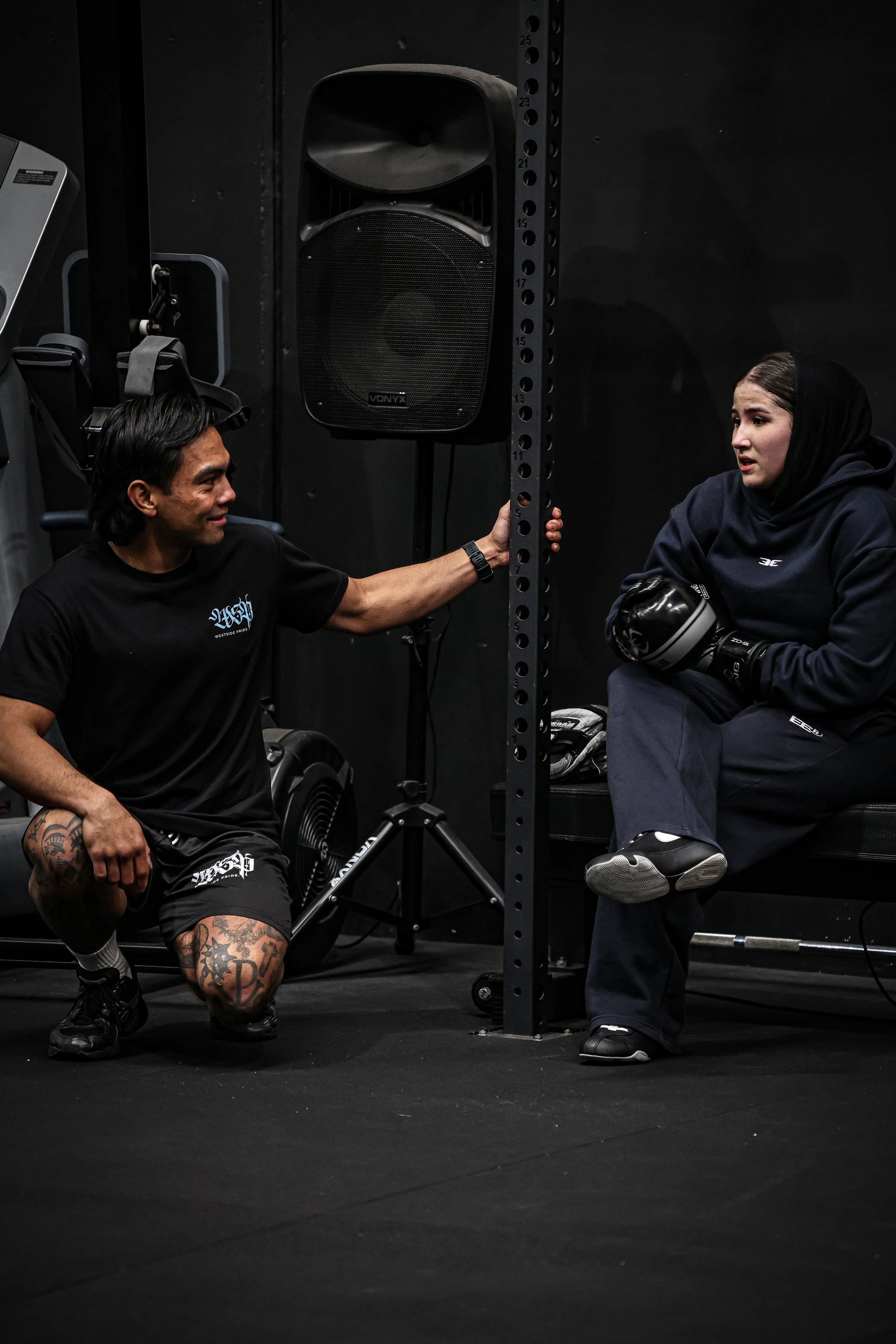 A man with tattoos on his arms and legs, wearing a black T-shirt and shorts, is squatting next to a black metal structure in a gym. He is holding a vertical pole and smiling. A woman wearing a black hoodie and black pants is sitting on a bench, holding boxing gloves and talking to him. A large speaker and gym equipment are visible in the background.
