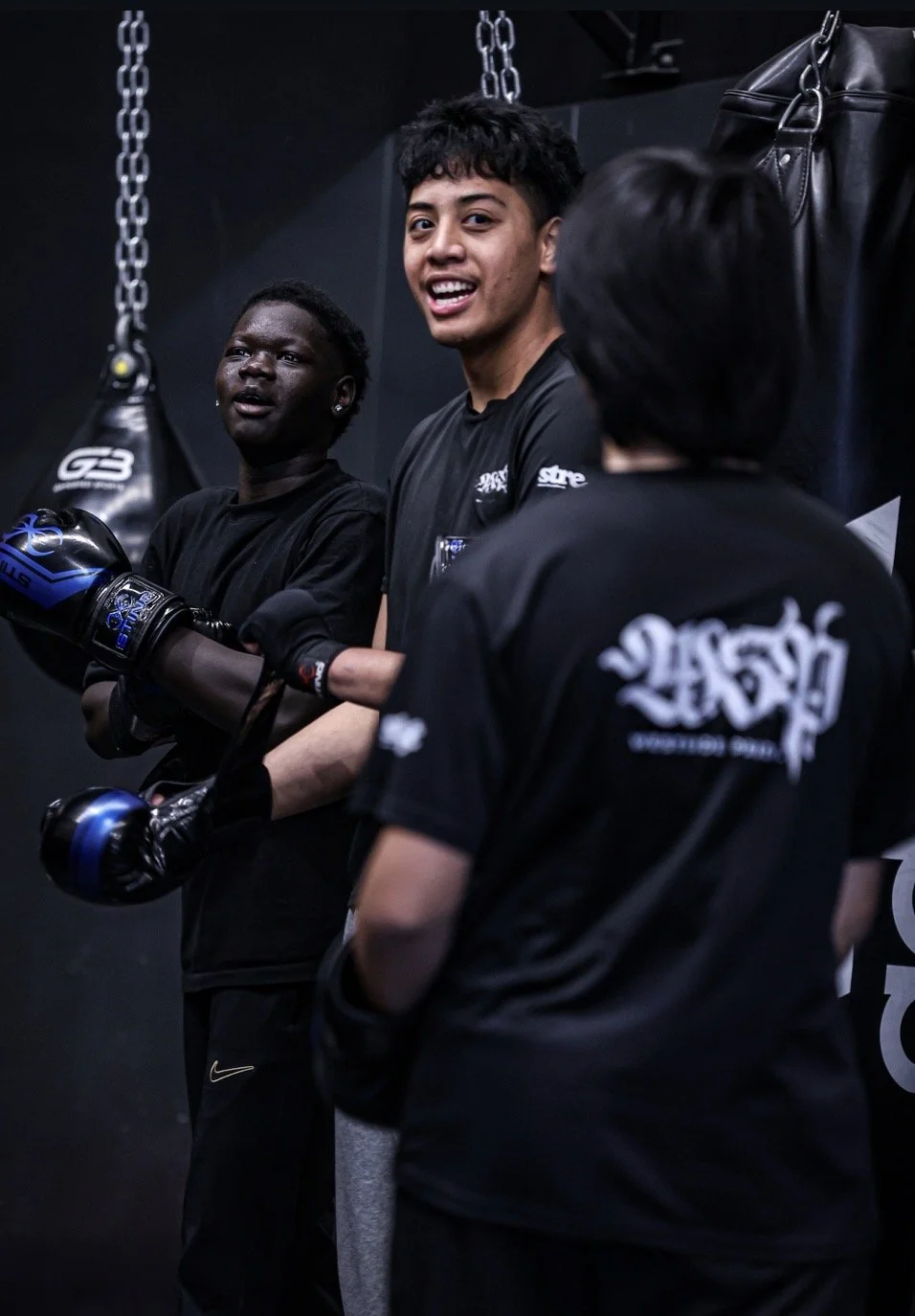 Three people in a gym, two of them wearing boxing gloves and black apparel, standing and conversing in front of hanging boxing bags.