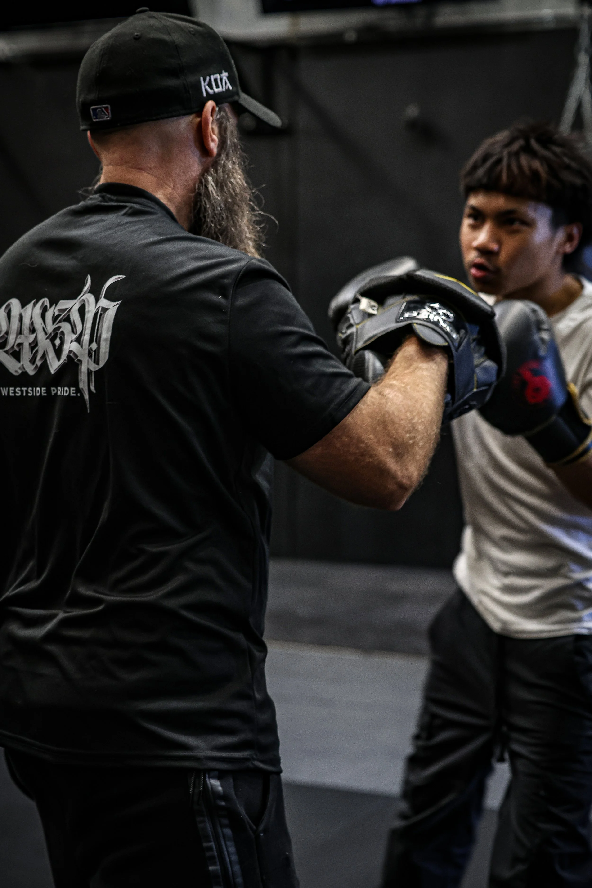 Image of a man with a beard wearing a black baseball cap and a black T-shirt coaching a young man in a white hoodie during a boxing training session, indoors with a dark background.