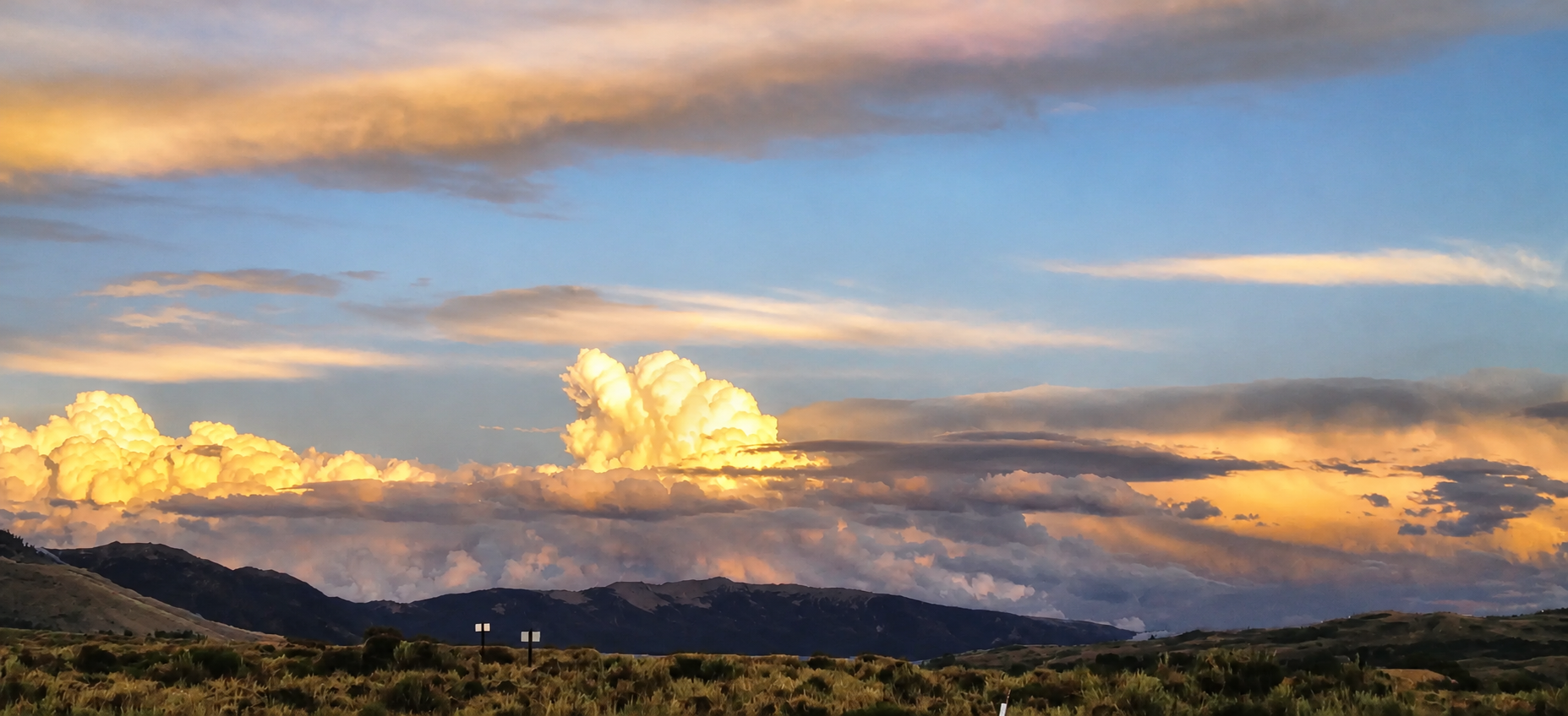 A landscape of cloudy skies at sunset over rolling hills with some bushes, orange and yellow clouds, and a patch of blue sky.