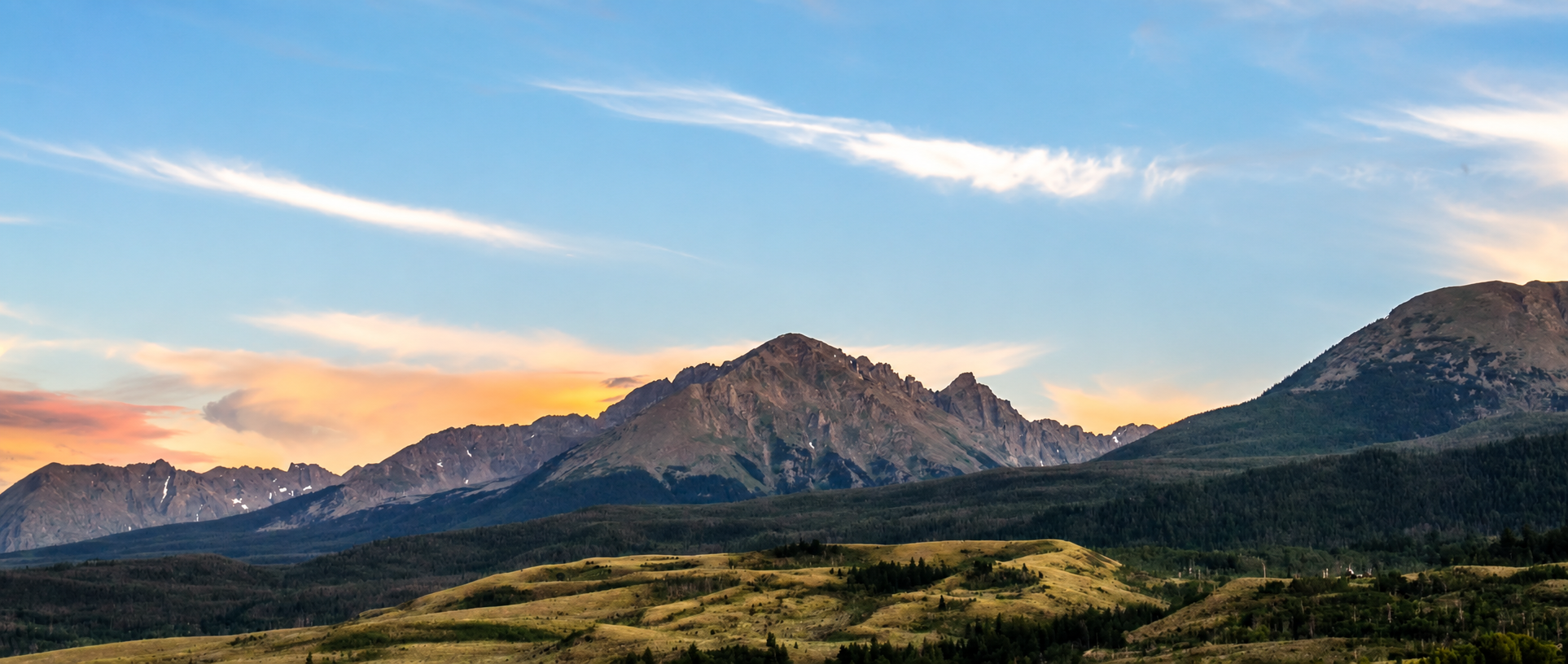 Mountains at sunset with green grassy hills in the foreground and clouds in a blue sky.