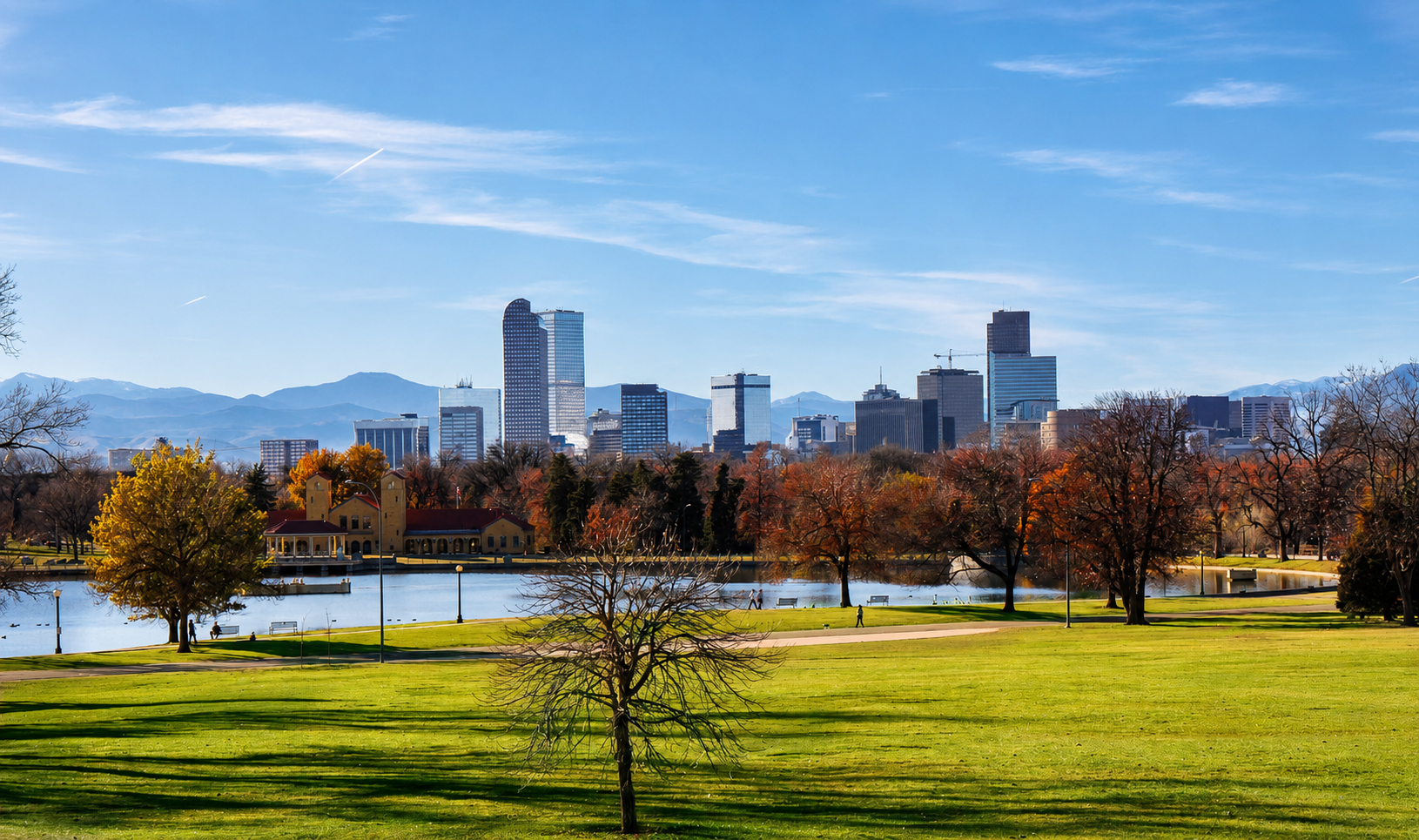 City skyline with tall buildings viewed from a park with a pond, green grass, trees with autumn leaves, and mountains in the background on a clear, sunny day.