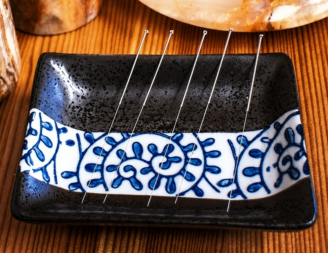 A black rectangular dish with a white and blue decorative pattern in the center, containing five stainless steel acupuncture needles.