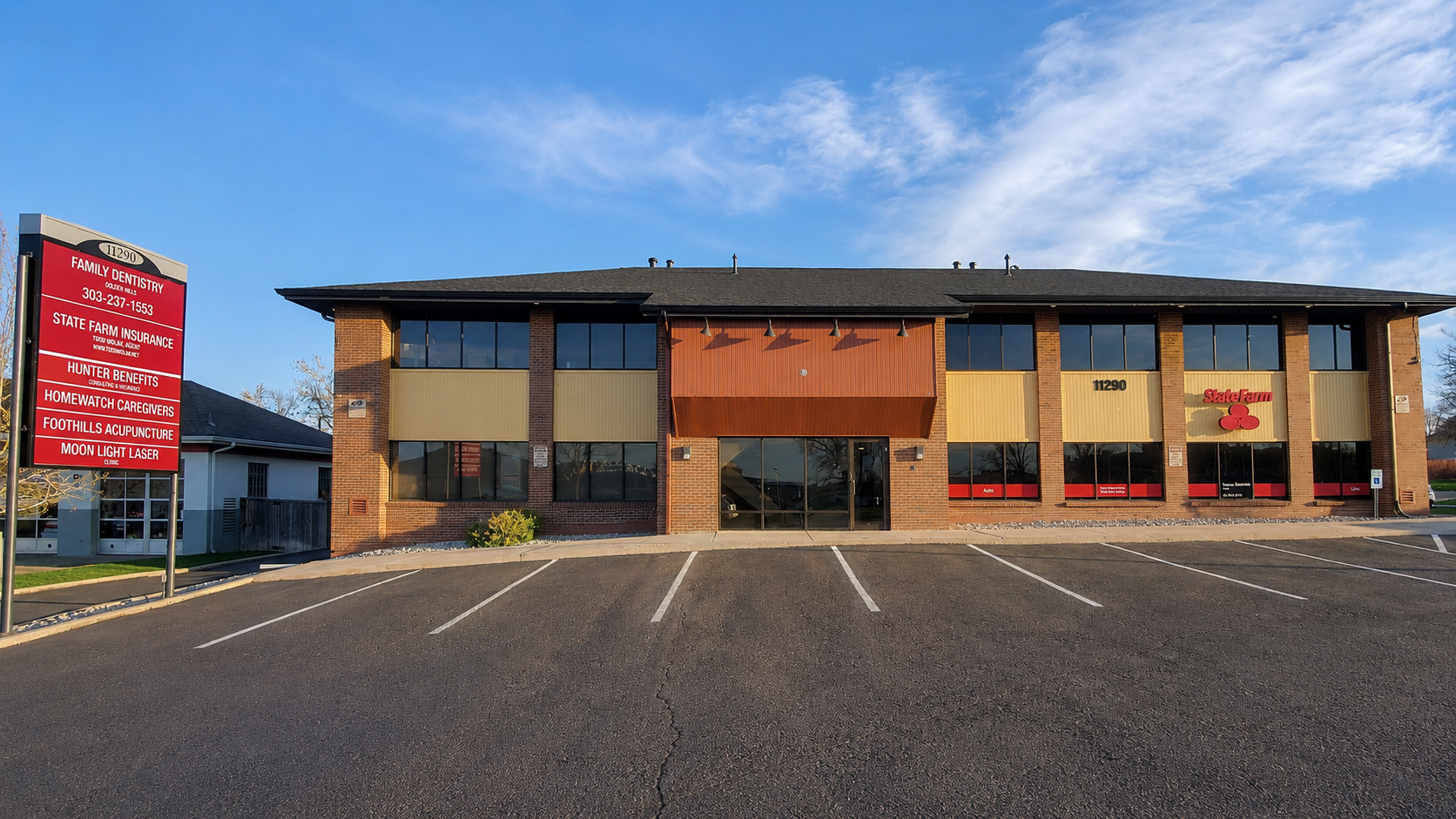 Front view of a brick commercial building with large windows, a parking lot with marked spaces, and a sign listing various health and insurance services in front.