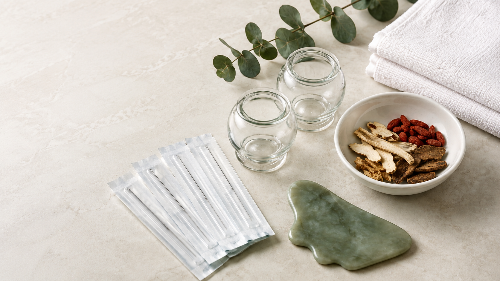 Relaxation setup with clear glasses, dried herbs and roots in a white bowl, white towels, a jade or green stone, and a sprig of greenery on a light-colored surface.