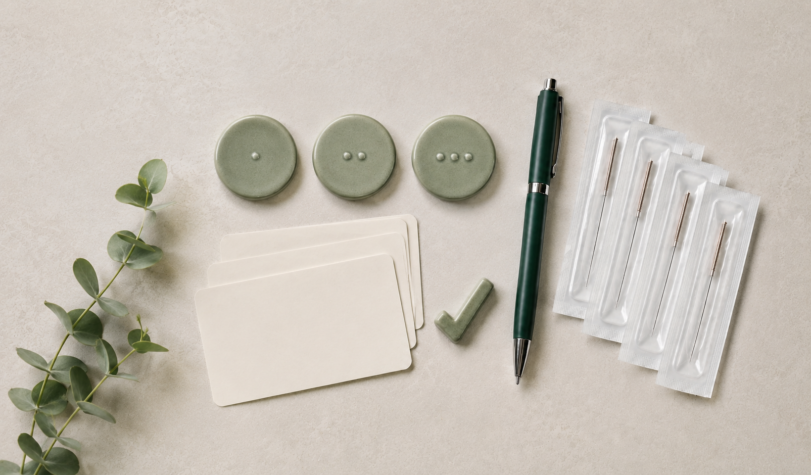 A collection of green buttons, blank white cards, a green pen, a small green block, a sprig of eucalyptus, and four individually packaged acupuncture needles on a light surface.