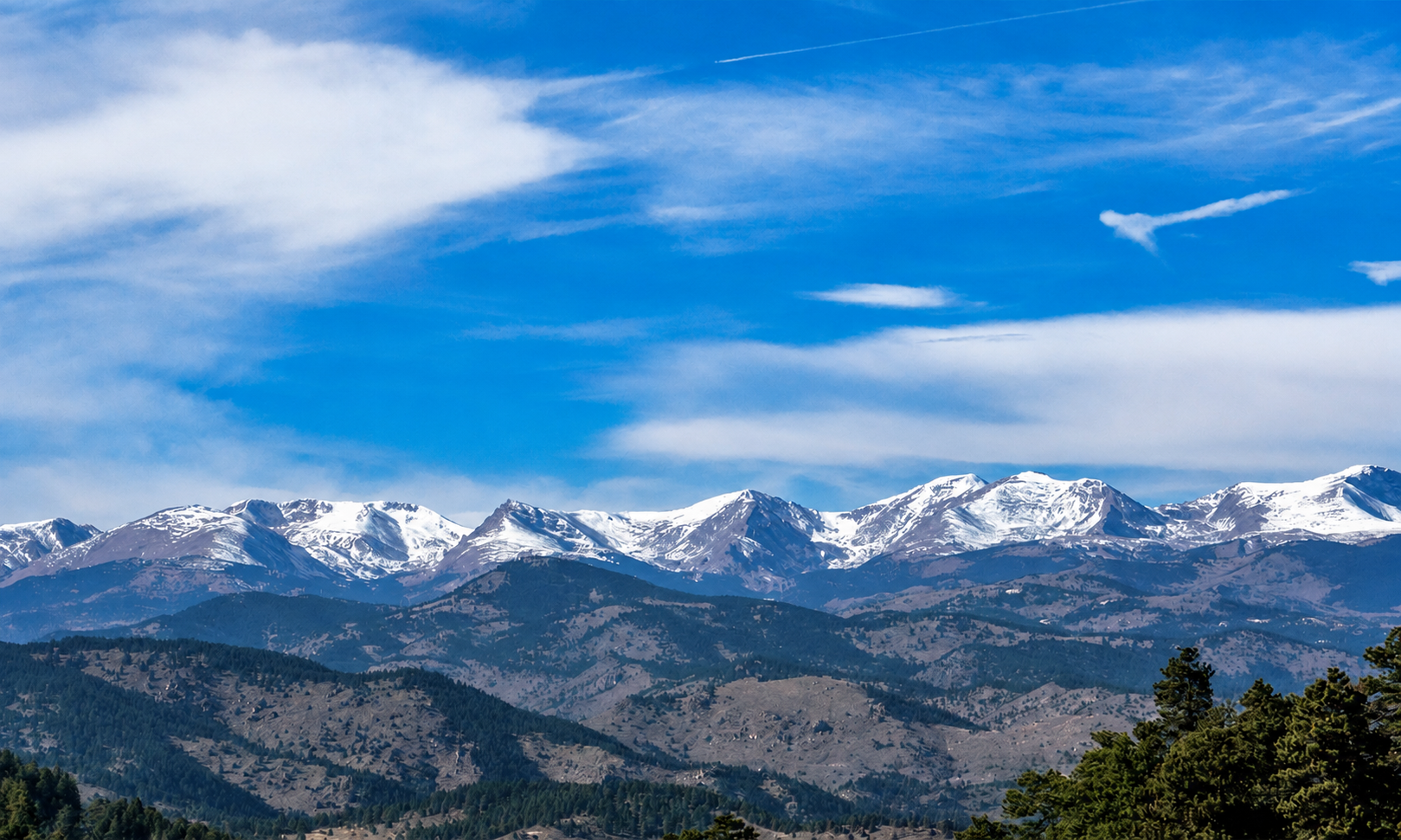 Snow-capped mountain range under a partly cloudy blue sky with trees in the foreground.
