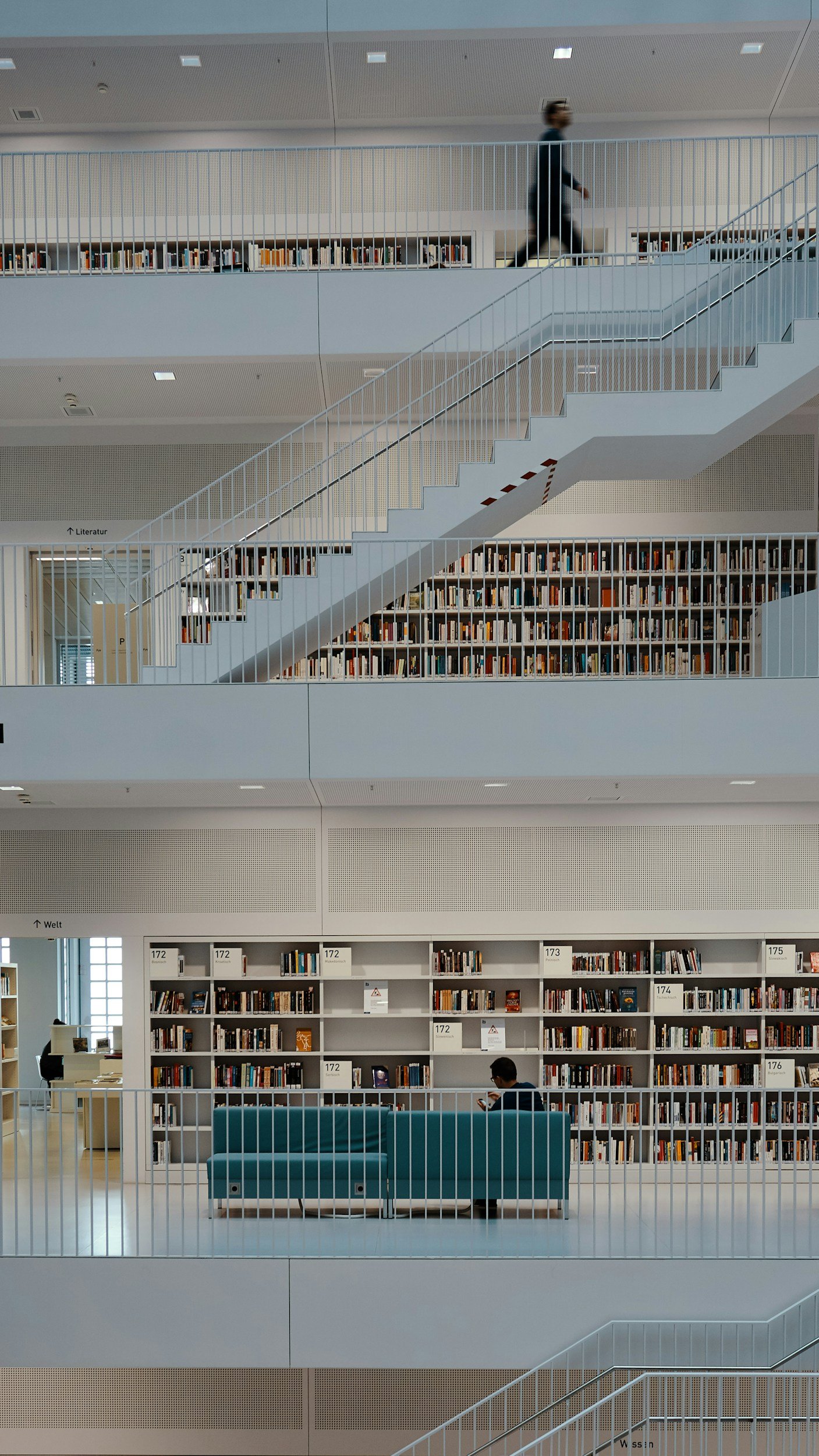 Interior of a modern library with multiple levels, white railings, and bookshelves filled with books. A person is walking on the upper level, and another person is sitting on a blue couch on the lower level, looking at a phone.