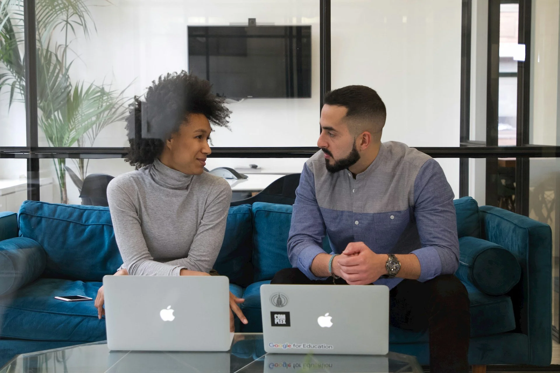 Two people sitting on a blue sofa in an office, engaged in conversation. There are two laptops with Apple logos on the table in front of them, and a large flat-screen TV mounted on the wall behind them.