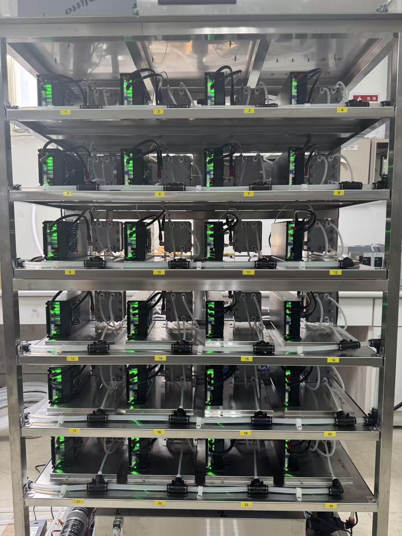 A metal rack holding multiple electronic devices connected with cables and glowing green lights, arranged on four shelves with yellow numbered labels.