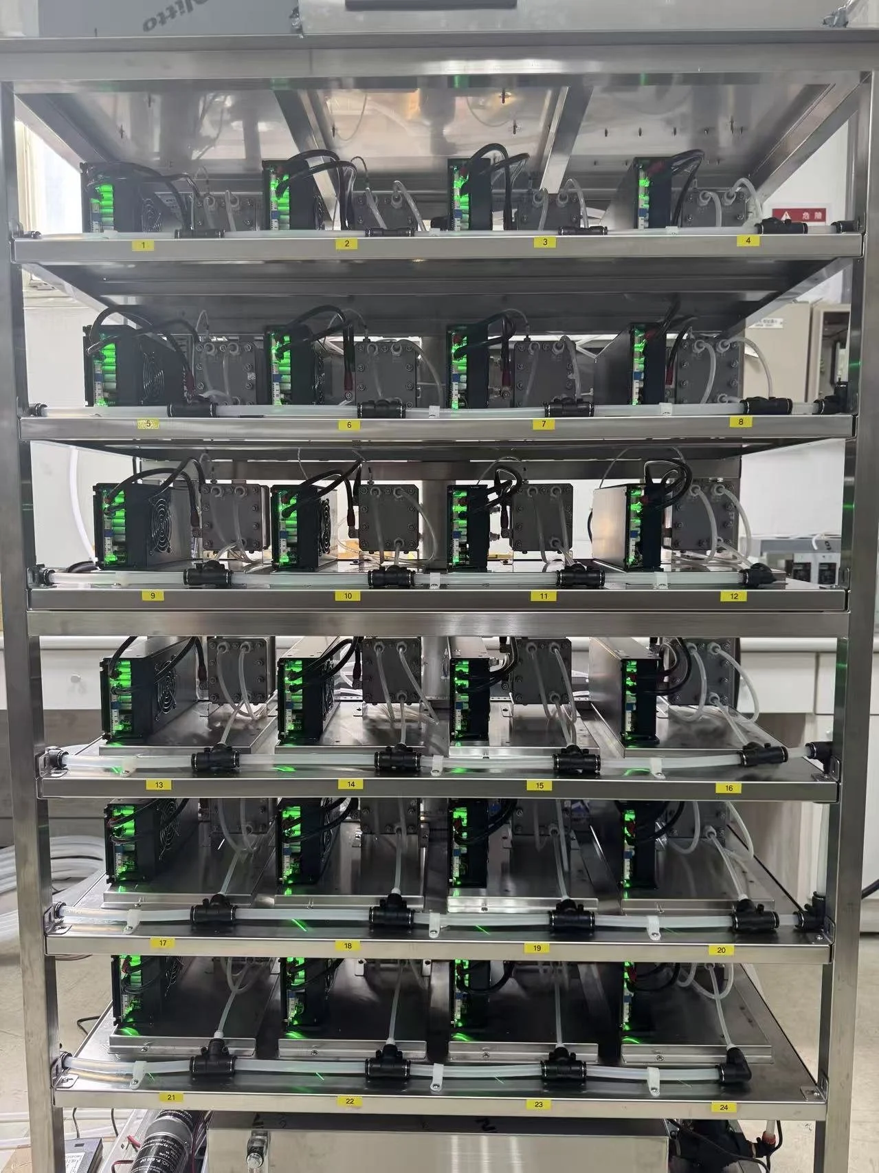 A metal rack holding multiple mining hardware units with green indicator lights, arranged in rows and columns, labeled with yellow tags numbered 1 to 24.