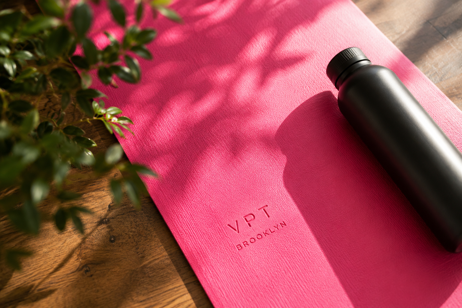 A black water bottle on a pink yoga mat with the text 'VPT Brooklyn' embossed on it, next to some green leaves on a wooden surface.