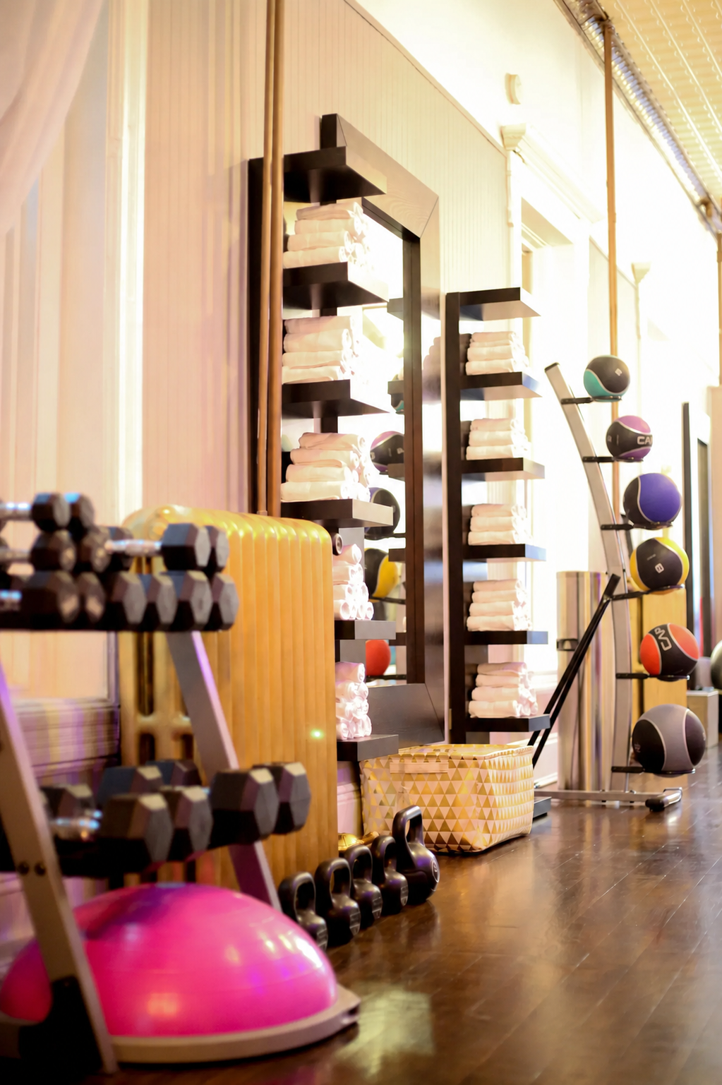 A sundrenched gym space with warm wood flooring. There is a rack with dumbbells, as well as a rack with medicine balls. There are wall mounted shelves and each shelf has a pyramid stack of white towels on them. Kettlebells are lined up on the floor.