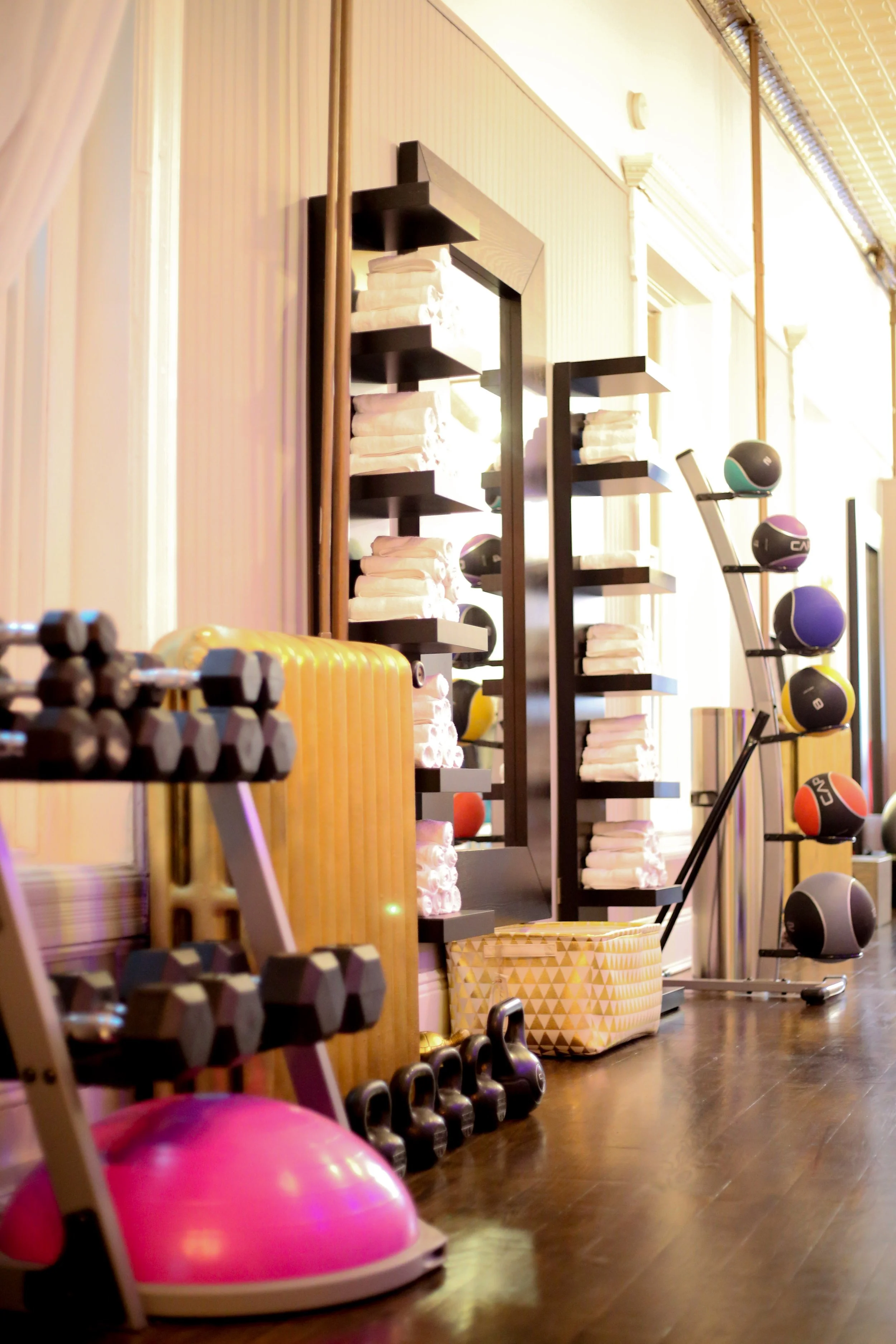 Gym or fitness studio equipment and accessories, including pink exercise balls, dumbbells, kettlebells, weight plates, and shelves with towels and medicine balls, in a well-lit room with wooden flooring.