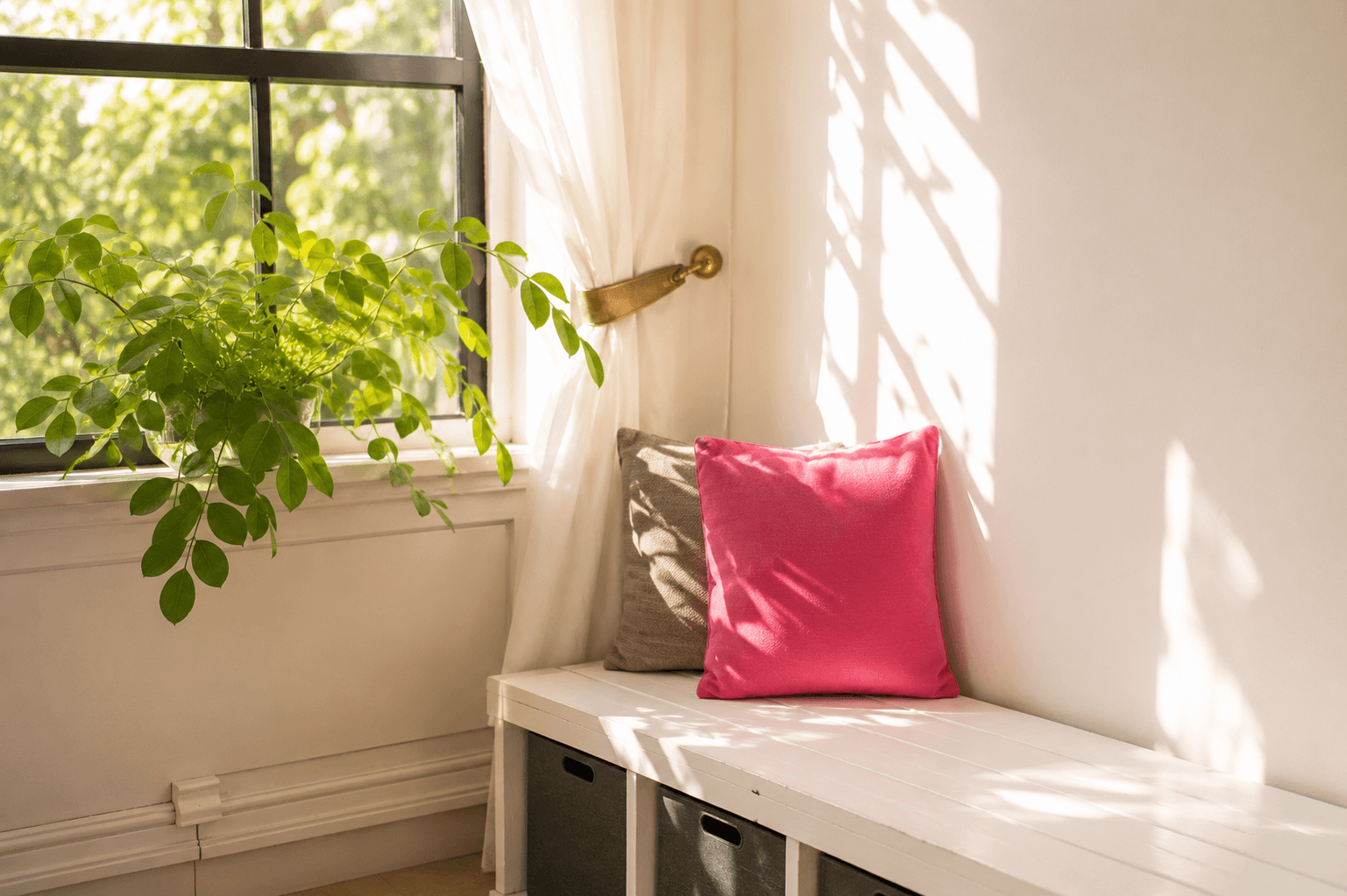 A bright window with white curtains, a green potted plant on the windowsill, a gray pillow, and a pink pillow on a white wooden bench inside a cozy, sunlit room.