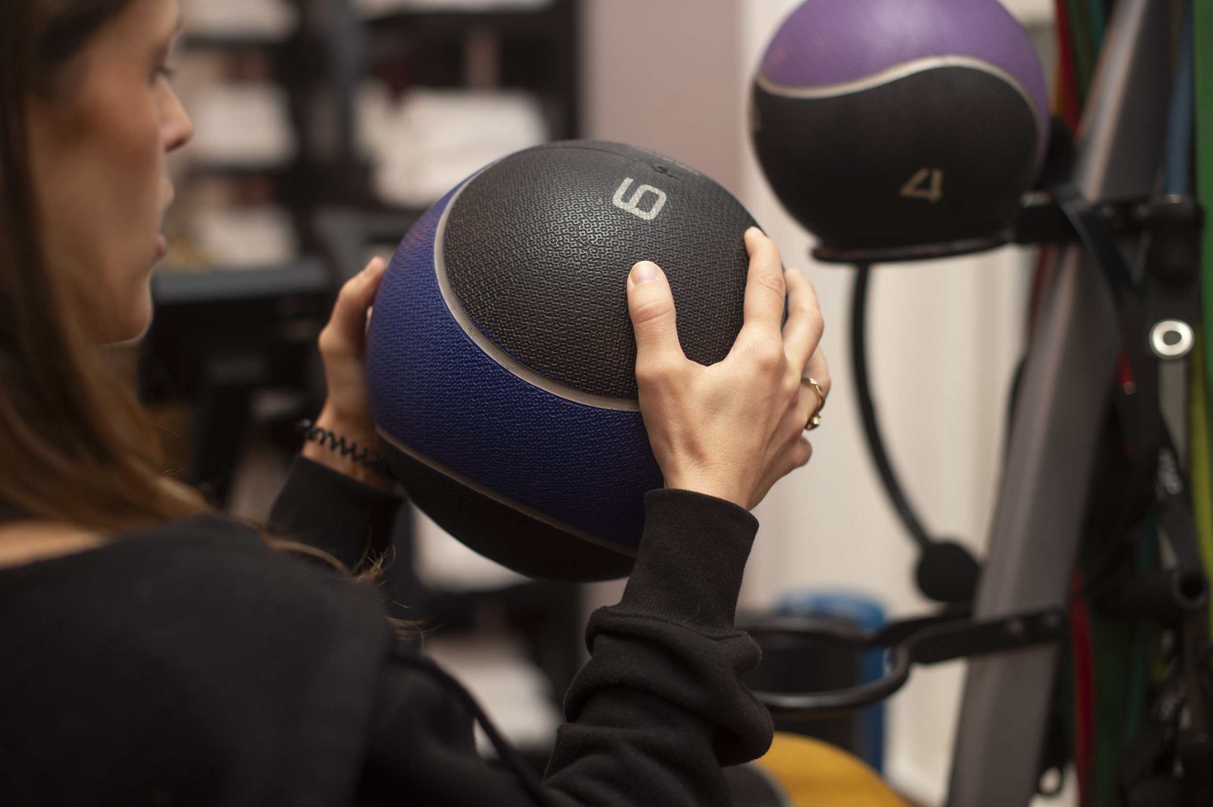 A woman in a black long sleeve shirt holding a black and blue medicine ball in a gym or fitness studio, with other colored medicine balls and fitness equipment visible in the background.