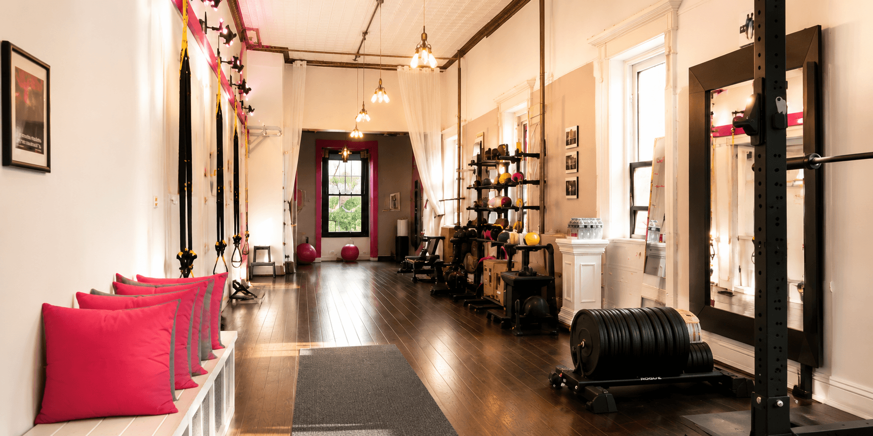 Bright fitness room with brick walls, hardwood floors, pink accents, and natural light from large windows. Equipment includes weights, resistance bands, a mirror, exercise balls, and a rowing machine.