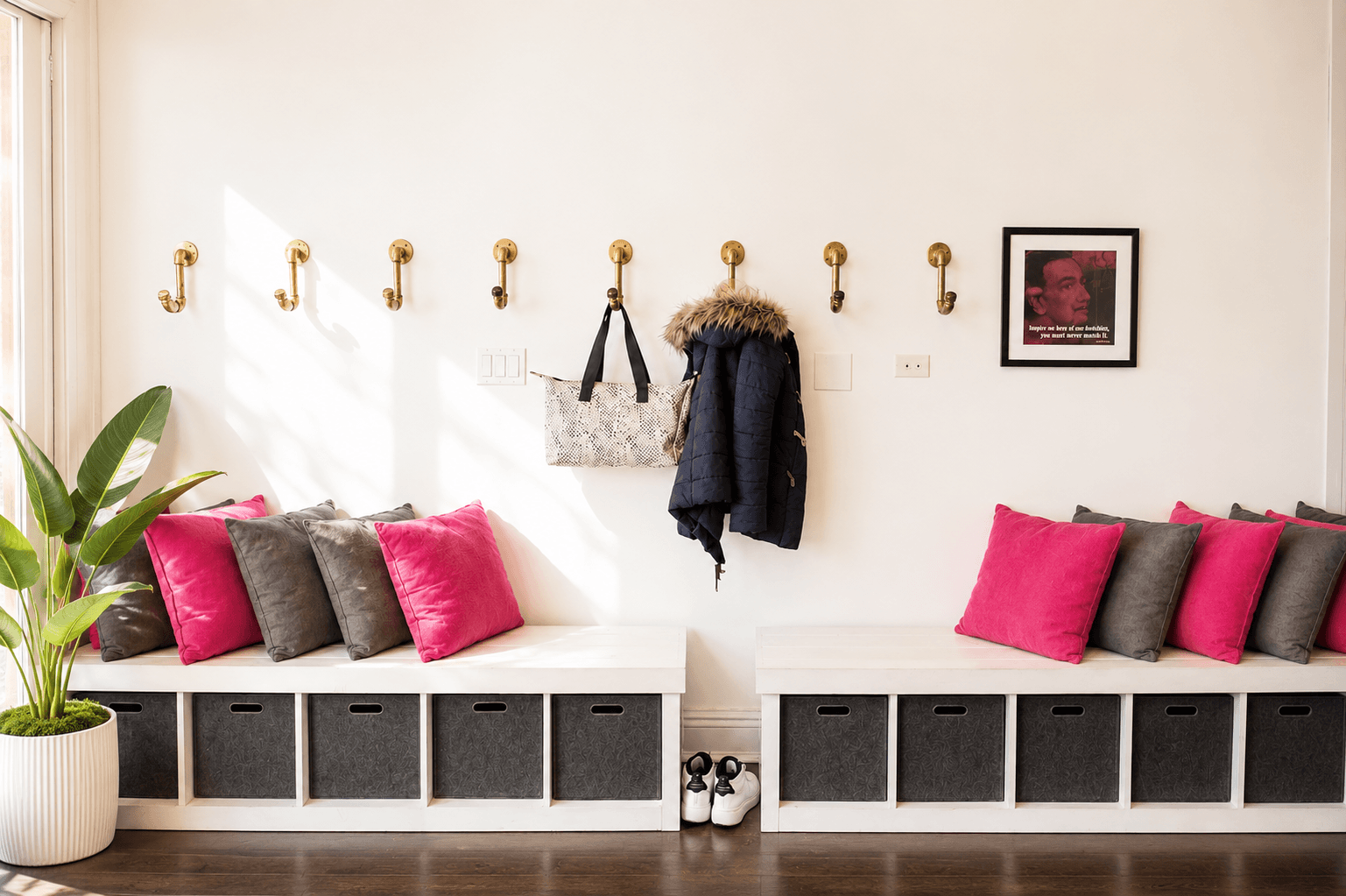 A mudroom with built-in white benches, pink and gray pillows, a row of brass hooks with coats and bags, a framed photo on the wall, and a potted plant sitting on the floor.