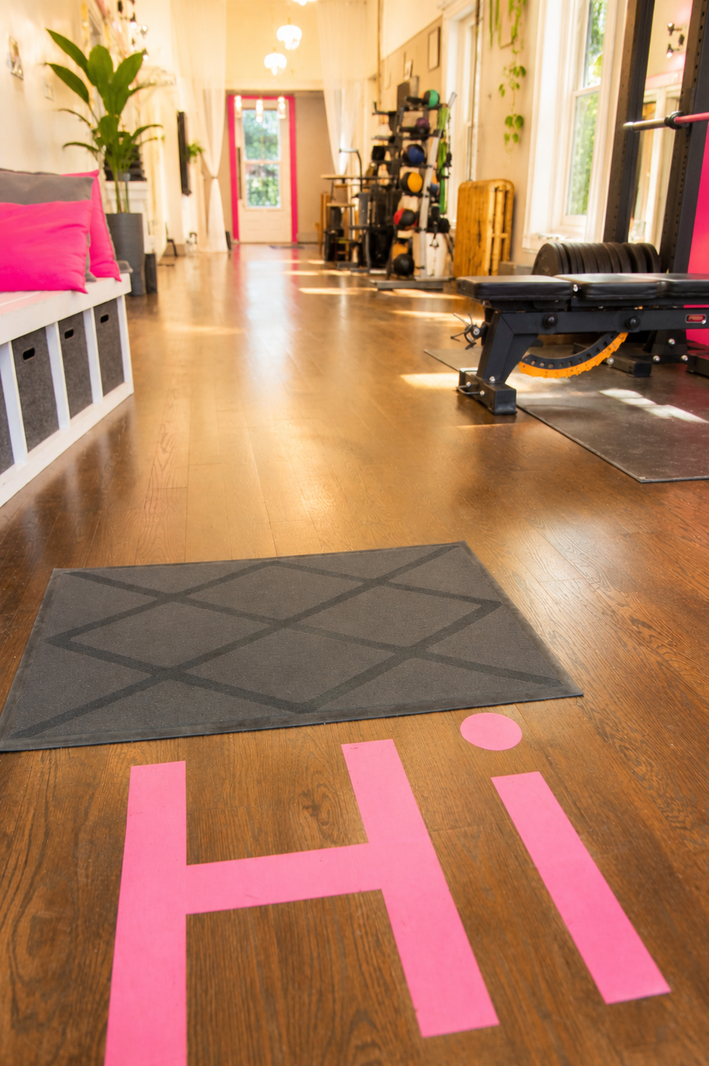 A gym with a wooden floor, a gray floor mat, and pink flooring letters spelling 'HI' in the foreground. The gym contains fitness equipment such as a weight bench, wall-mounted weights, and wall-mounted shelves with various fitness gear. Large windows let in natural light, and there are green plants along the walls.