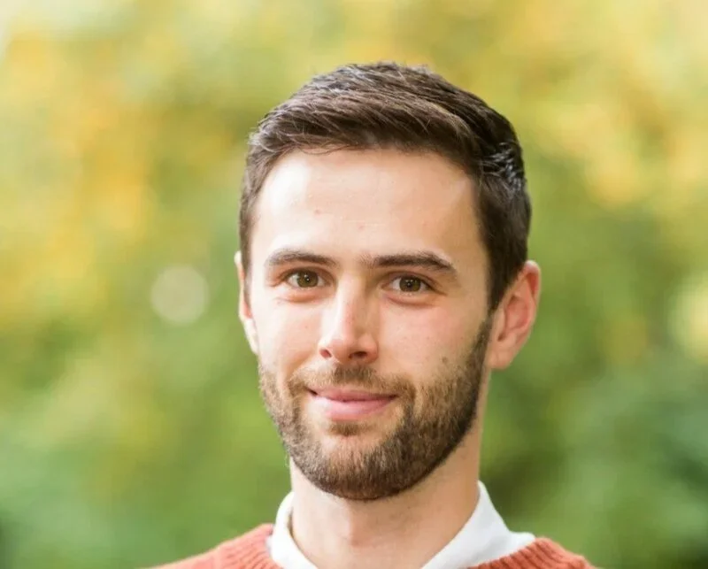 Portrait of a young man with short brown hair and a beard, smiling outdoors with trees in the background.