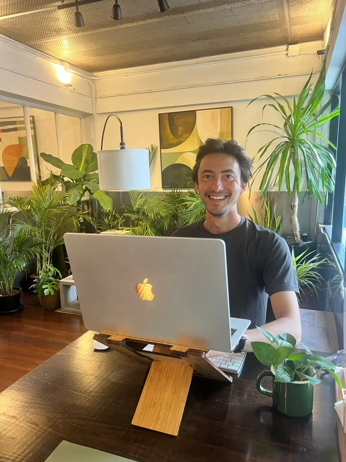 A smiling man sitting at a wooden table in a room filled with green plants, using a MacBook laptop on a wooden stand.
