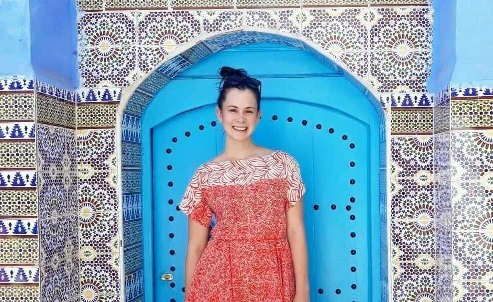 Young woman smiling in front of a bright blue arched door, surrounded by Moroccan-style colorful tiled walls.