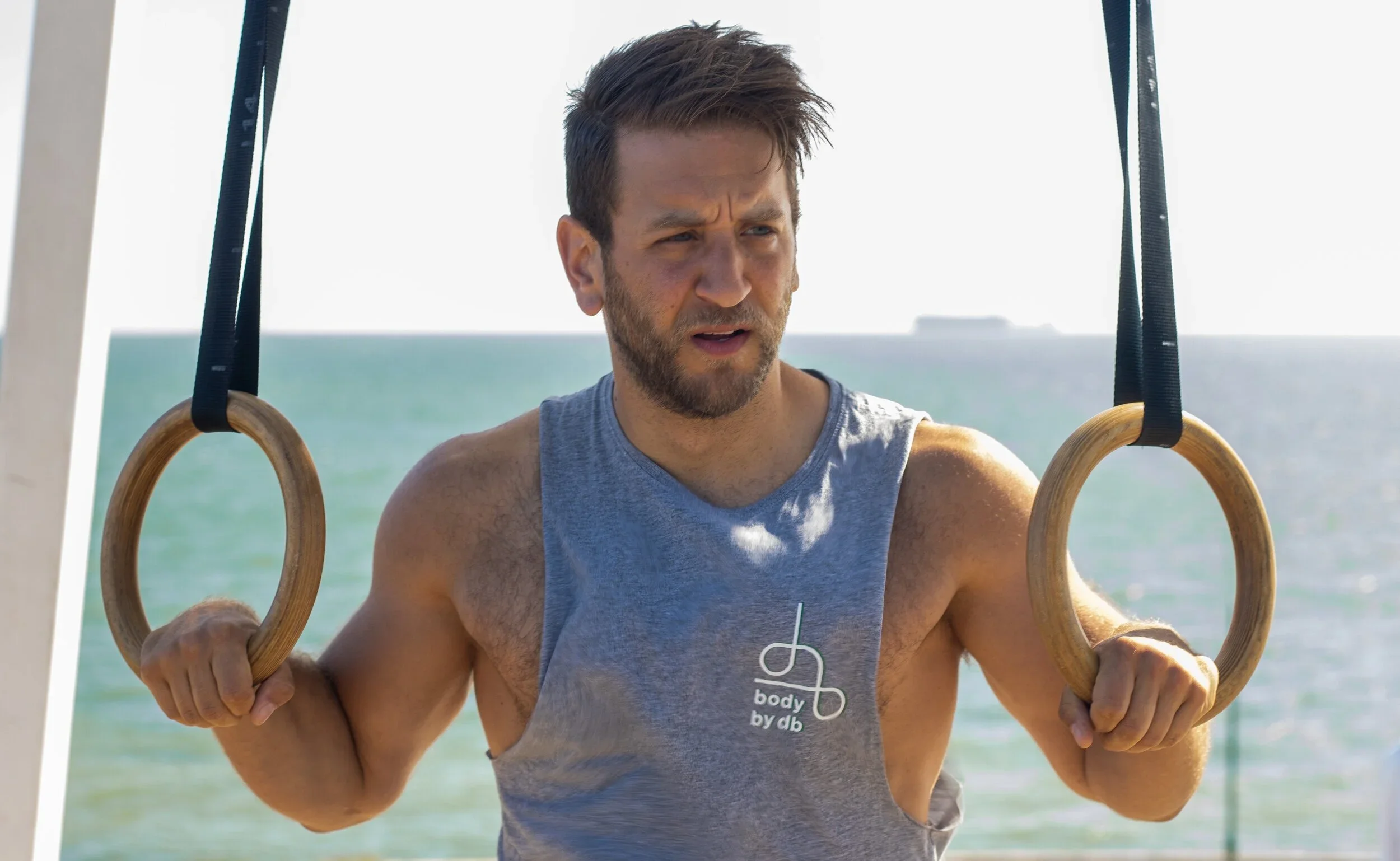 A man with brown hair and a beard performing a workout on gymnastic rings outdoors near the ocean, wearing a gray sleeveless shirt and holding the rings with both hands.