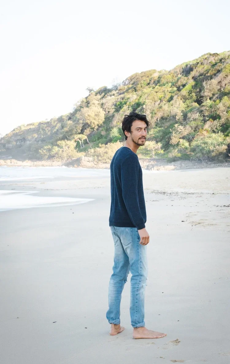 A young man with dark hair and a goatee standing barefoot on a sandy beach near a hill covered with green trees, looking back at the camera.