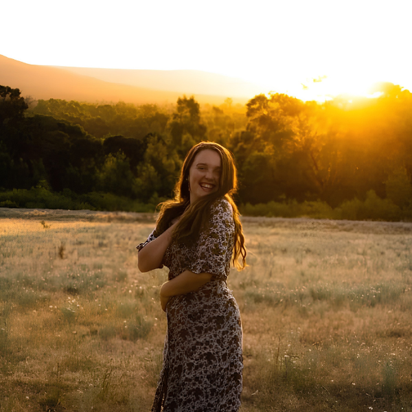 A woman with long hair wearing a floral dress standing in an open field at sunset, smiling and looking at the camera.