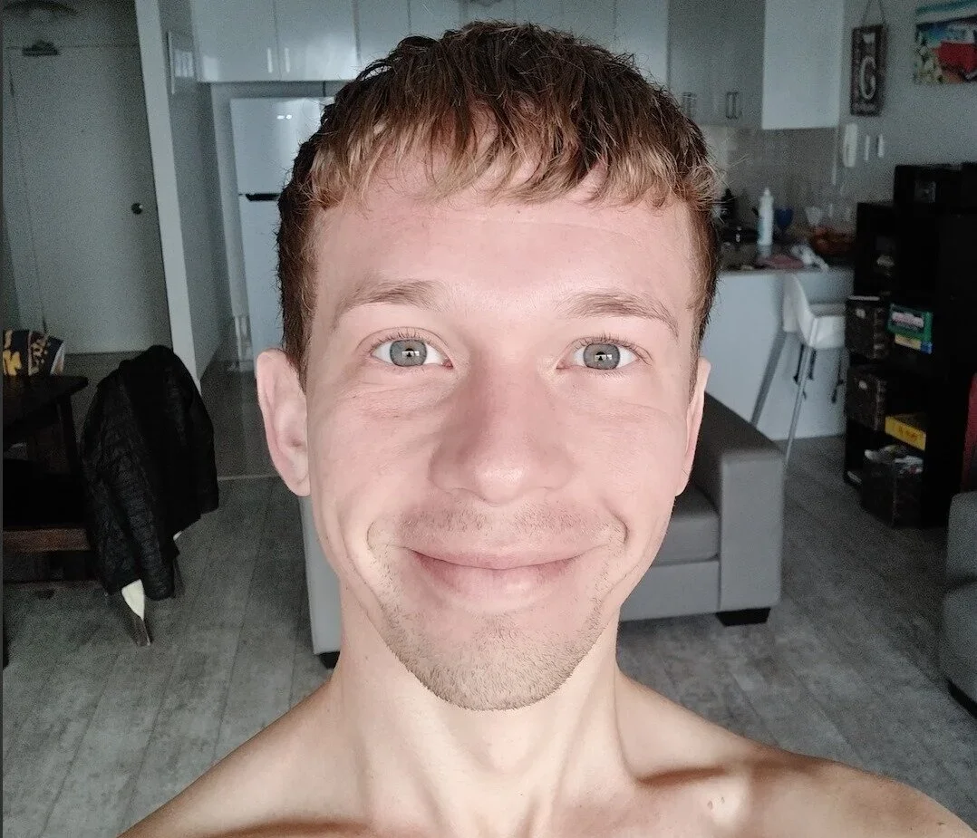 A young man with short brown hair and blue eyes smiling at the camera indoors.
