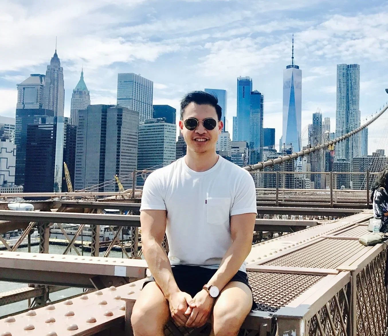 A man wearing sunglasses and a white T-shirt sitting on the Brooklyn Bridge with the New York City skyline in the background.