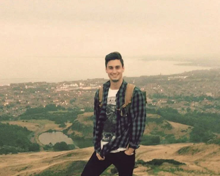 A young man with dark hair, wearing a plaid shirt and white t-shirt, stands on a hilltop with a backpack, smiling at the camera. Behind him is a scenic landscape with water, greenery, and a distant city skyline under a cloudy sky.