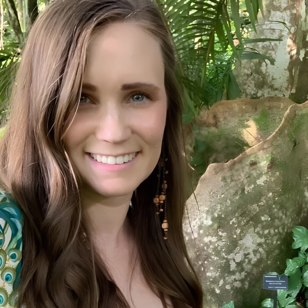 A young woman with long brown hair, smiling, in a lush, green jungle or botanical garden setting.