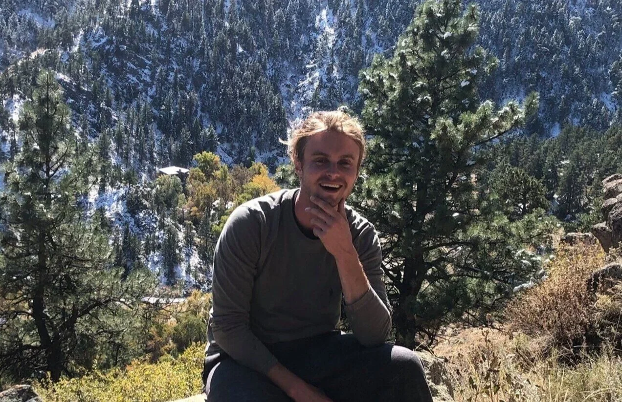 A young man with blond hair sitting outdoors on a rocky surface surrounded by pine trees and mountains in the background, smiling and touching his chin.
