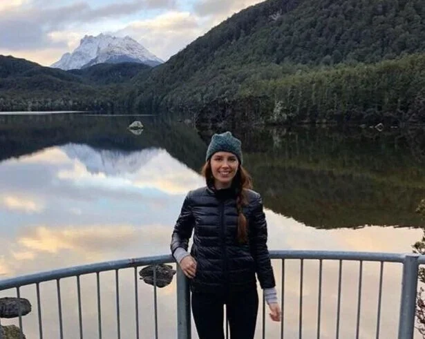 A woman standing by a metal railing near a lake with a mountain and forest in the background during daytime.