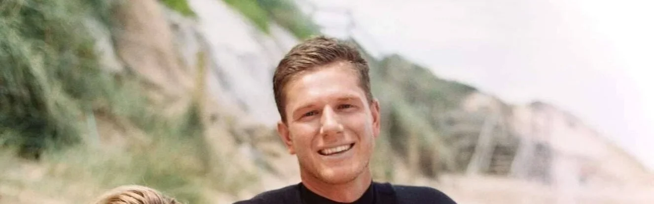 Smiling young man with short light brown hair outdoors near a rocky hill.