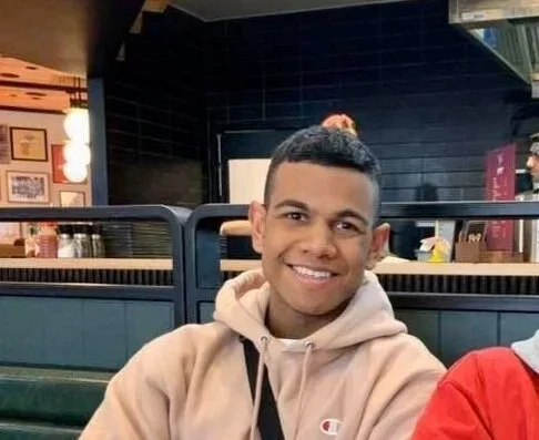 Smiling young man with short dark hair wearing a beige hoodie sitting in a restaurant or cafe.